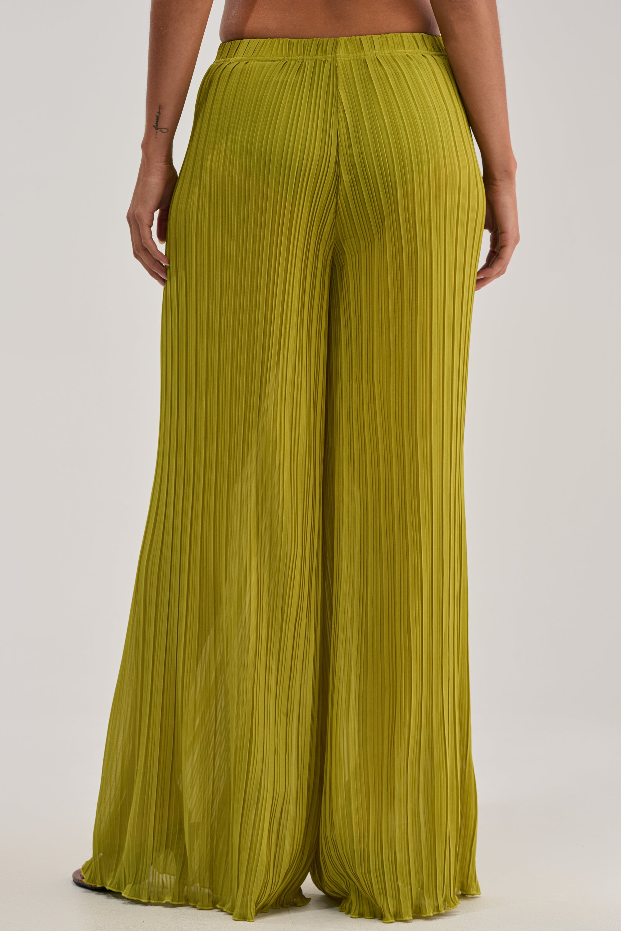 A person shown from the waist down wears the CRUSHIN ON YOU PALAZZO PANT IN CHARTREUSE—sheer, pleated chiffon pants that stand out against a plain, light background, making them bold resort wear essentials.