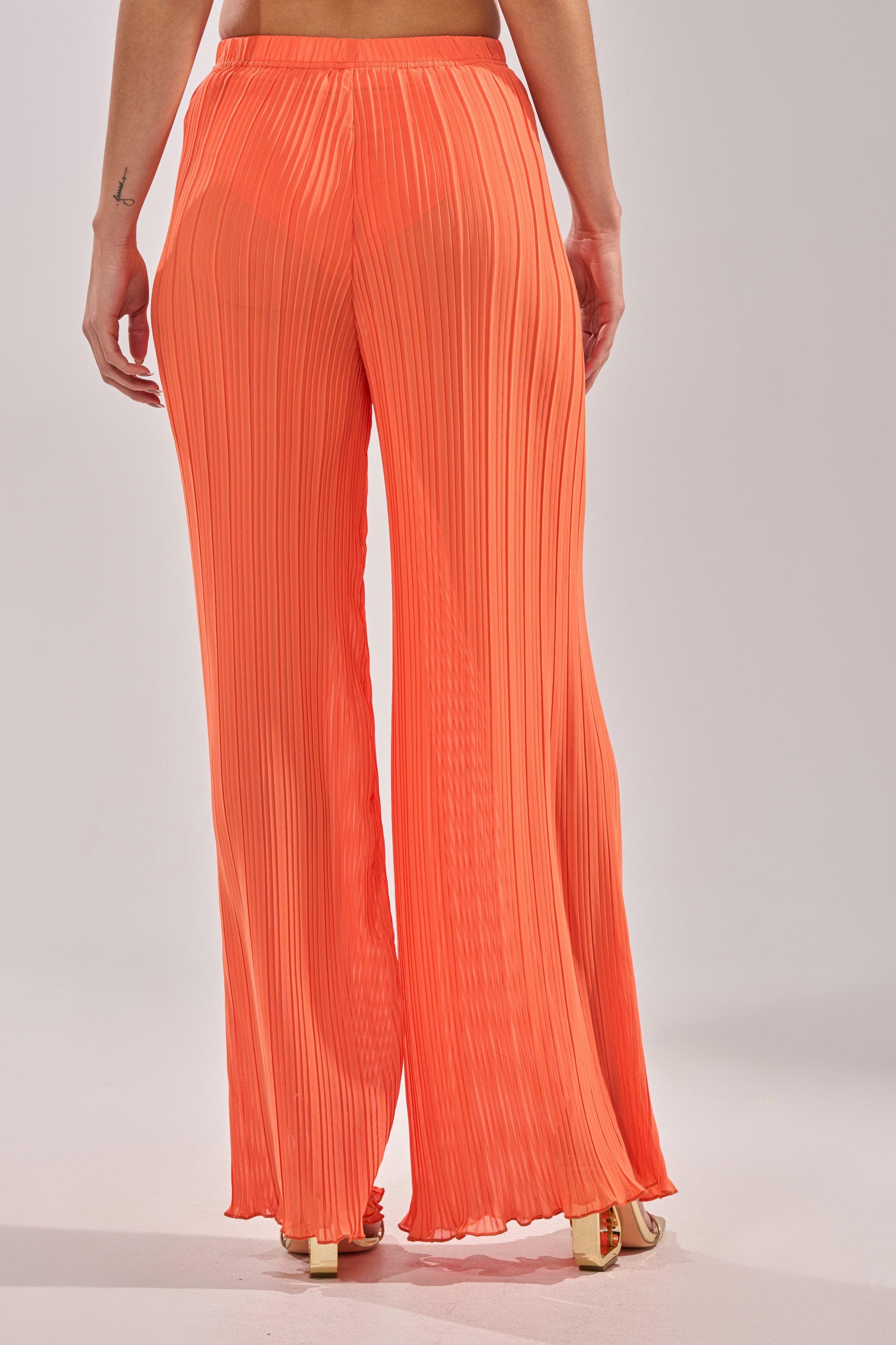 A person stands on a light background, viewed from behind, wearing the CRUSHIN ON YOU PALAZZO PANT—high-waisted, wide-leg orange pants paired with beige heels—an effortlessly chic piece for any vacation wardrobe. 5