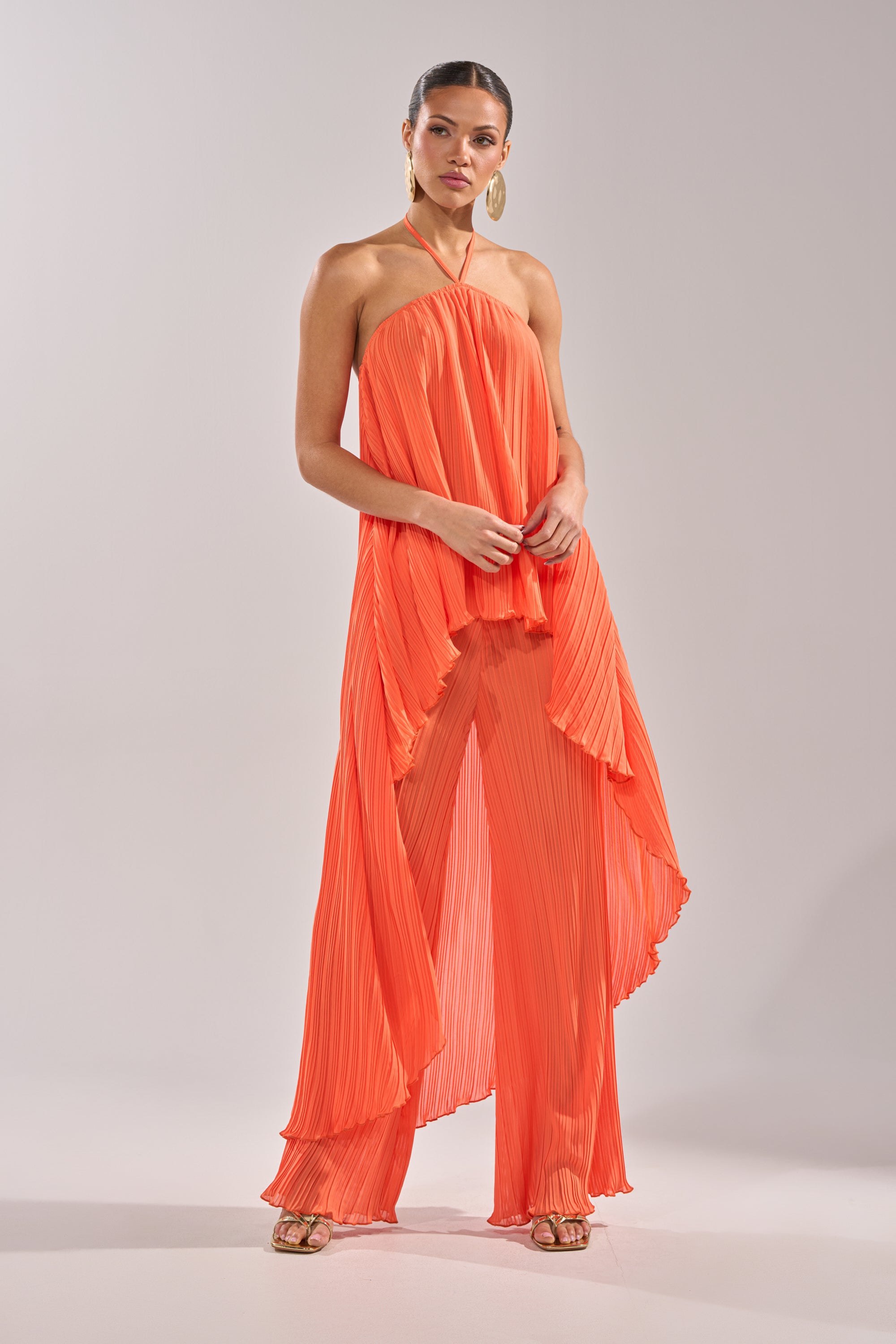 A woman poses confidently in the CRUSHIN ON YOU PALAZZO PANT, a bright orange halter-neck outfit with flowing fabric and wide-leg pants—ideal for a chic vacation look. She pairs it with gold heels, hoop earrings, slicked-back hair, and natural makeup. 7
