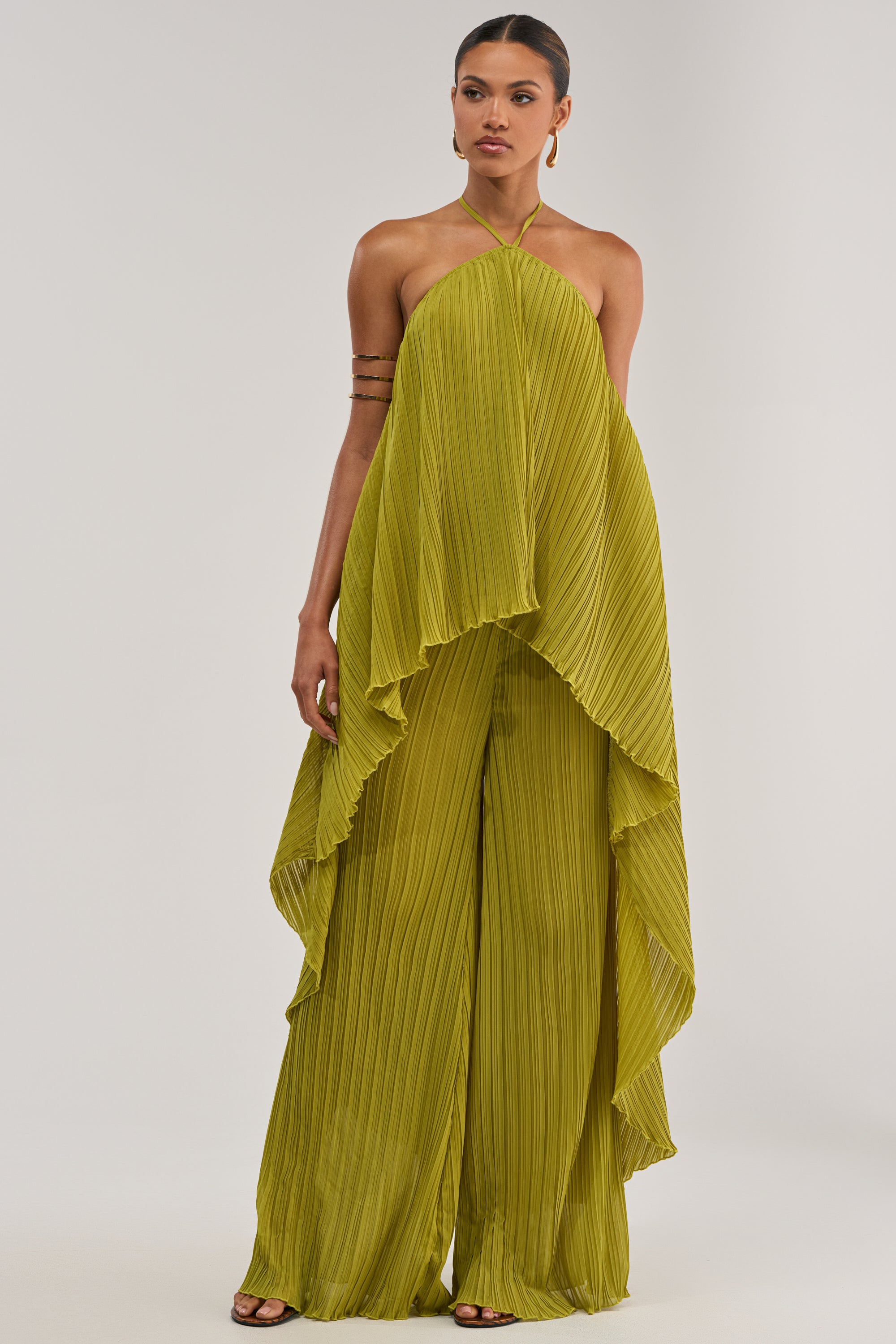 A woman models the CRUSHIN ON YOU BLOUSE IN CHARTREUSE, a loose-fitting, pleated top with a halter neckline. Draped, asymmetrical layers evoke resort style, accented by hoop earrings and arm cuffs against a plain background.