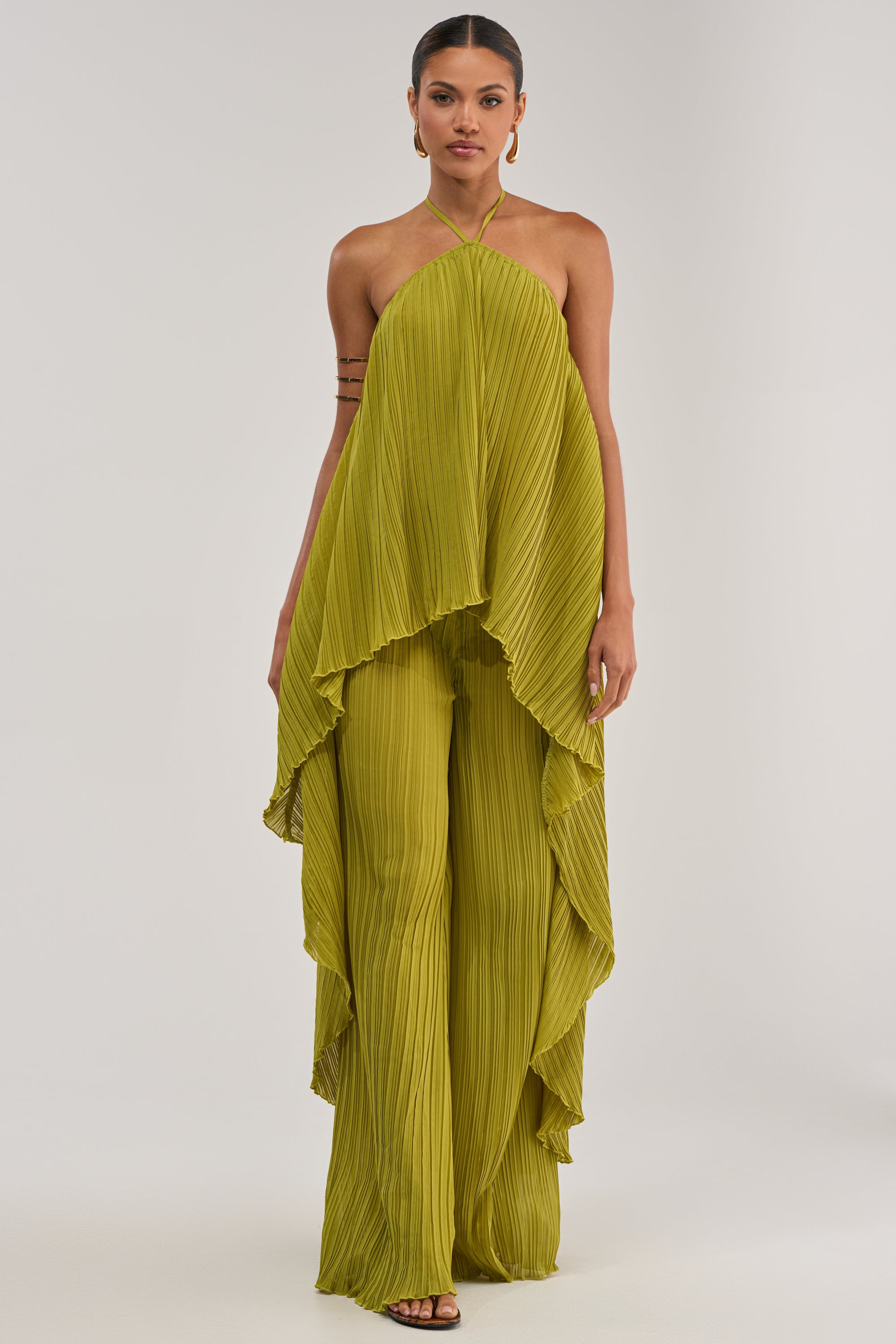 A person wears the CRUSHIN ON YOU BLOUSE IN CHARTREUSE, a flowing pleated chiffon top with a high-low hem, paired with matching wide-leg pants—an effortlessly chic look for modern resort wear.