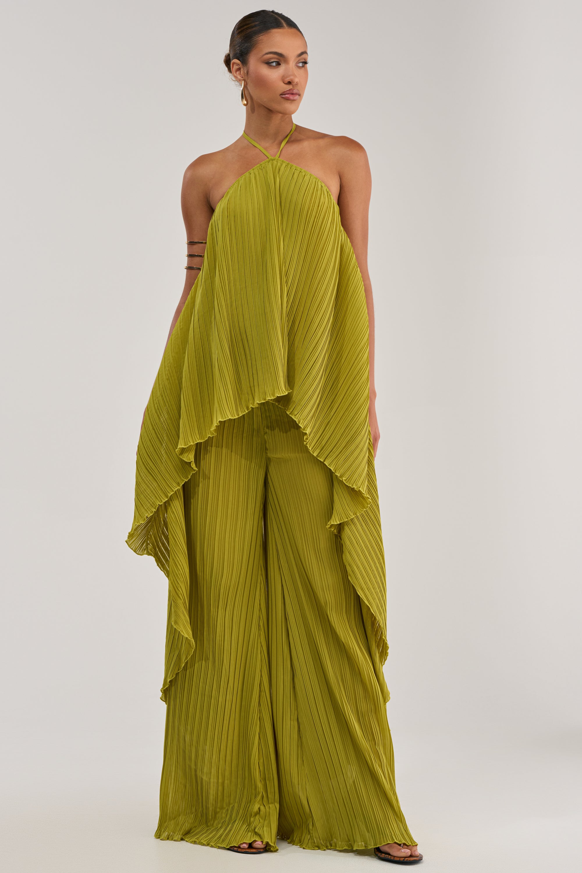 A woman wears the CRUSHIN ON YOU BLOUSE IN CHARTREUSE, featuring pleats and a flowing, layered design reminiscent of resort wear, paired with wide-leg pants, standing against a plain white background.