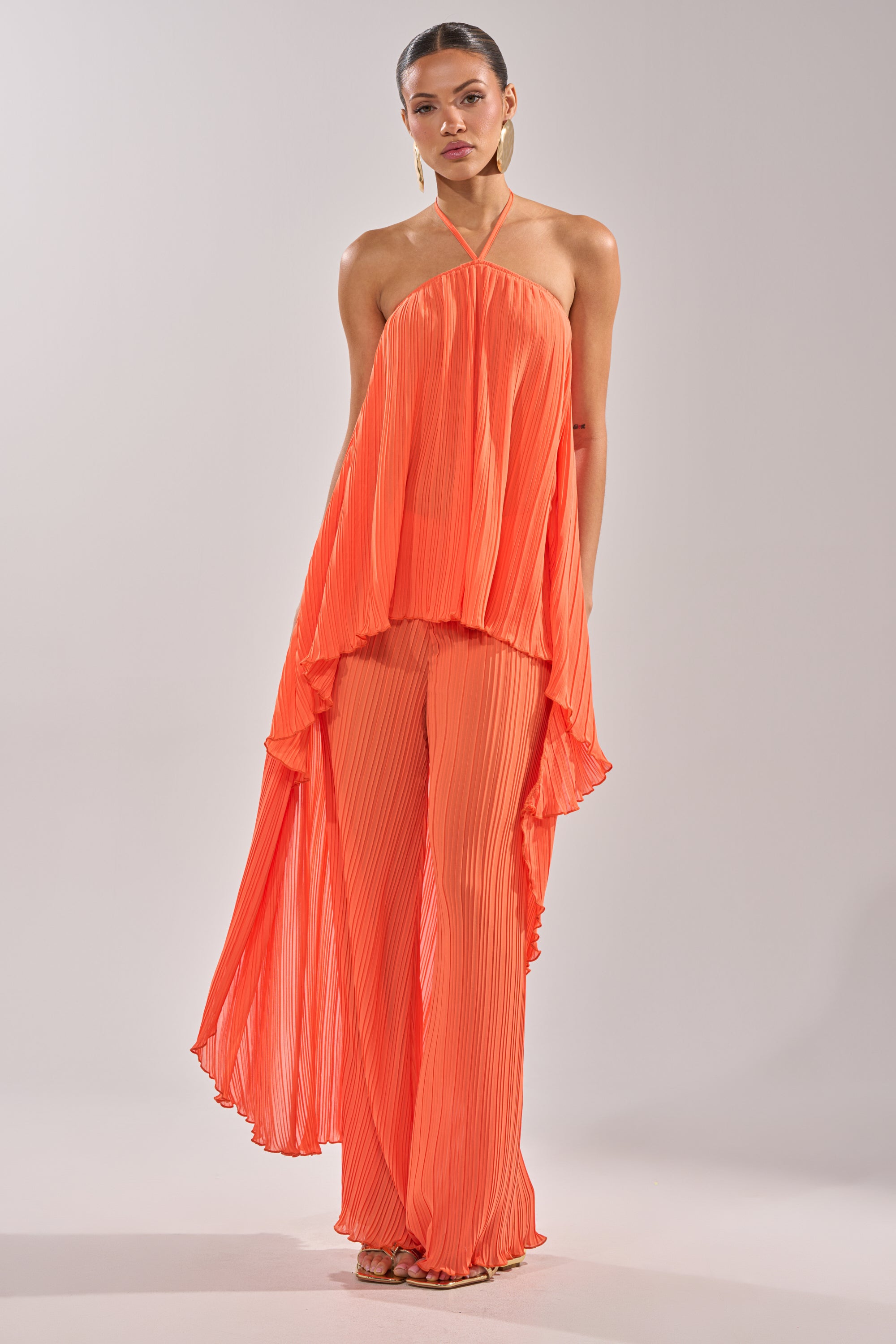 A woman wears the CRUSHIN ON YOU BLOUSE, a flowing orange halter top with pleats and an asymmetrical hem, paired with matching wide-leg pants. Her hair is pulled back and hoop earrings complete her chic, resort-ready style against a plain background. 8