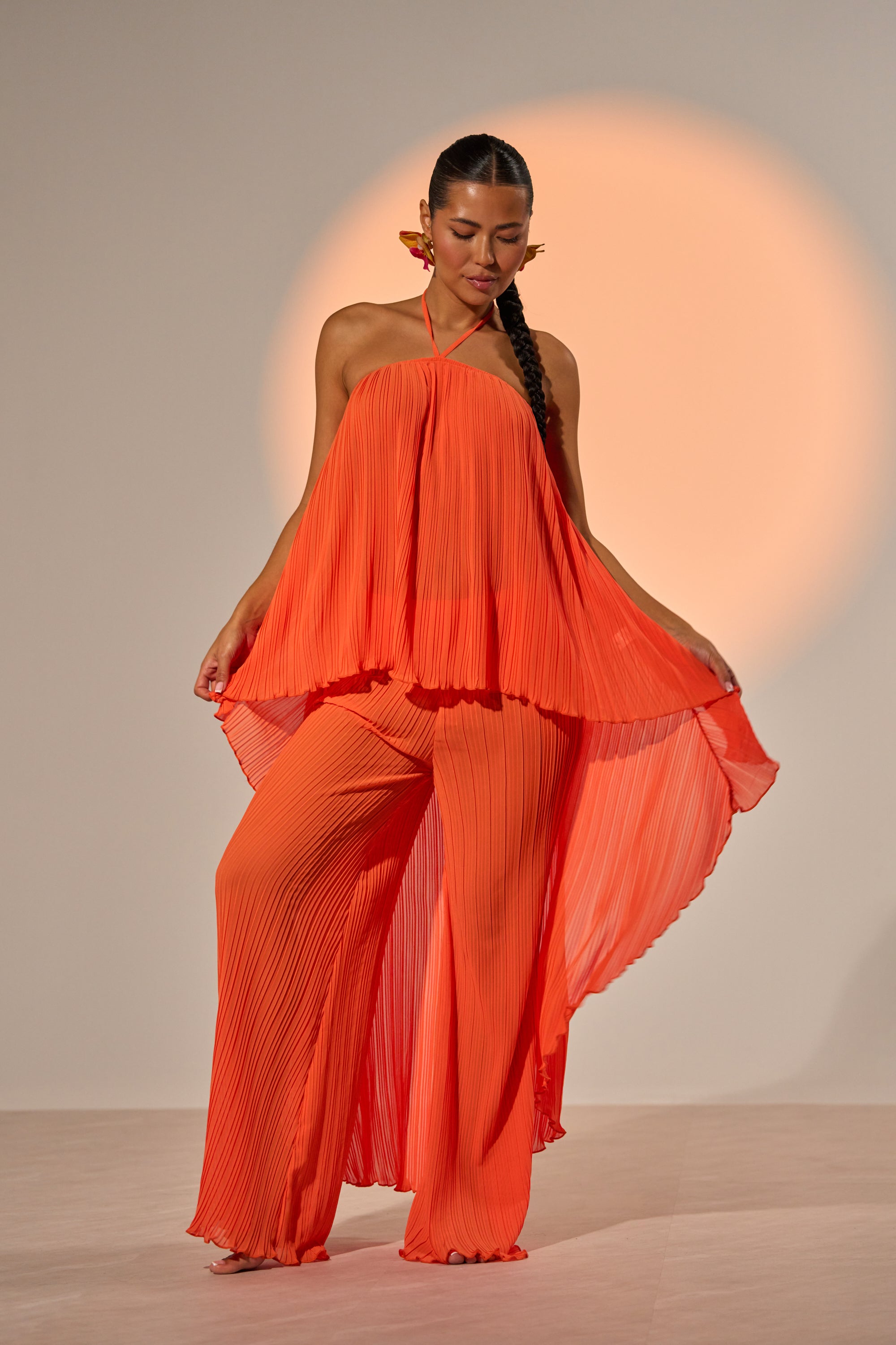 A woman stands gracefully against a neutral backdrop, modeling the CRUSHIN ON YOU BLOUSE in sunset orange with a pleated asymmetrical hem, paired with matching wide-leg pants and highlighted by a soft, circular orange light. 0