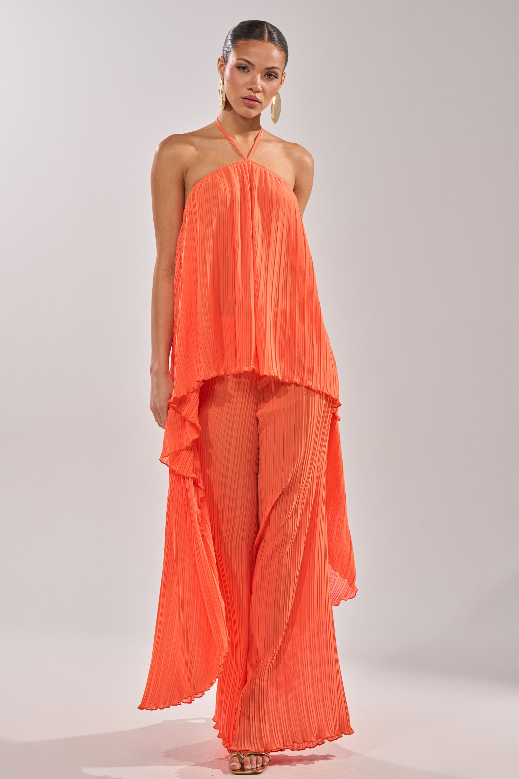 A woman wears the CRUSHIN ON YOU BLOUSE in sunset orange, featuring pleated flowy fabric and an asymmetrical hem, paired with matching wide-leg pants. She finishes the look with hoop earrings, heeled sandals, and her hair pulled back. 4
