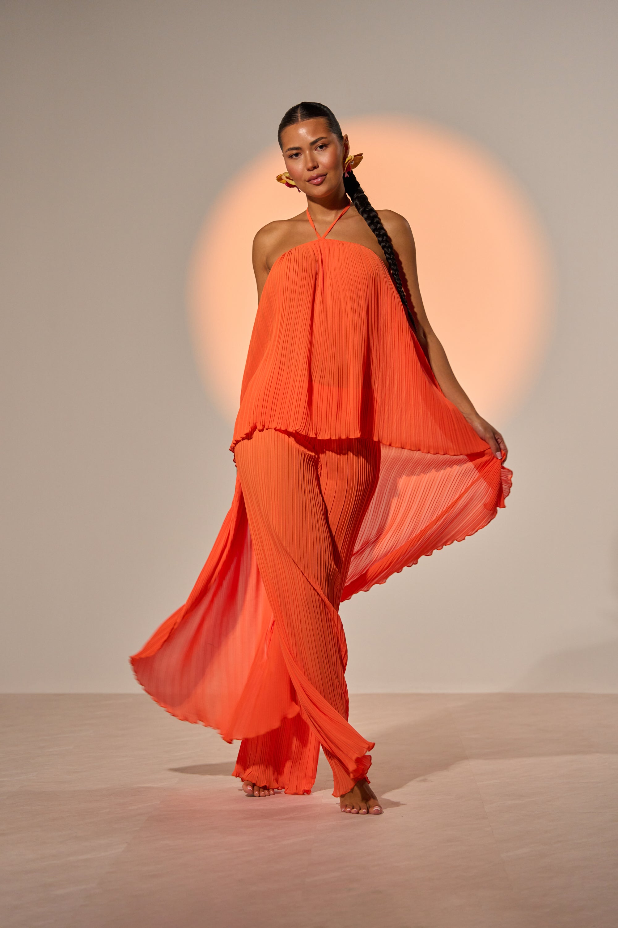 A woman with long braided hair models the CRUSHIN ON YOU BLOUSE in sunset orange, paired with matching wide-leg pants and barefoot, standing against a neutral backdrop with a soft orange glow behind her. 1