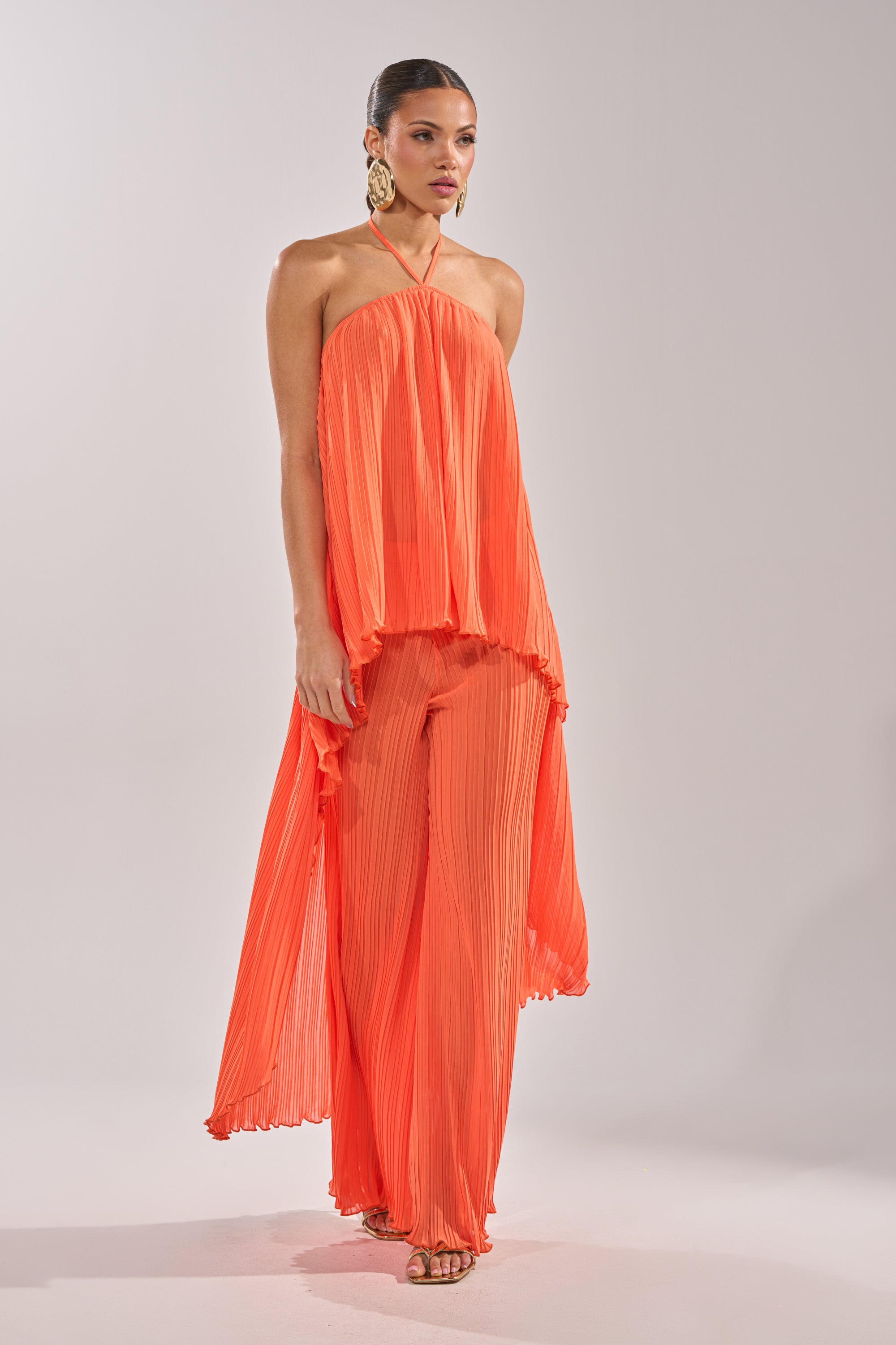 A woman wears the CRUSHIN ON YOU BLOUSE in sunset orange, featuring a halter neckline and pleated high-low hem, with matching wide-leg pants, gold statement earrings, and heels against a plain light background. 5