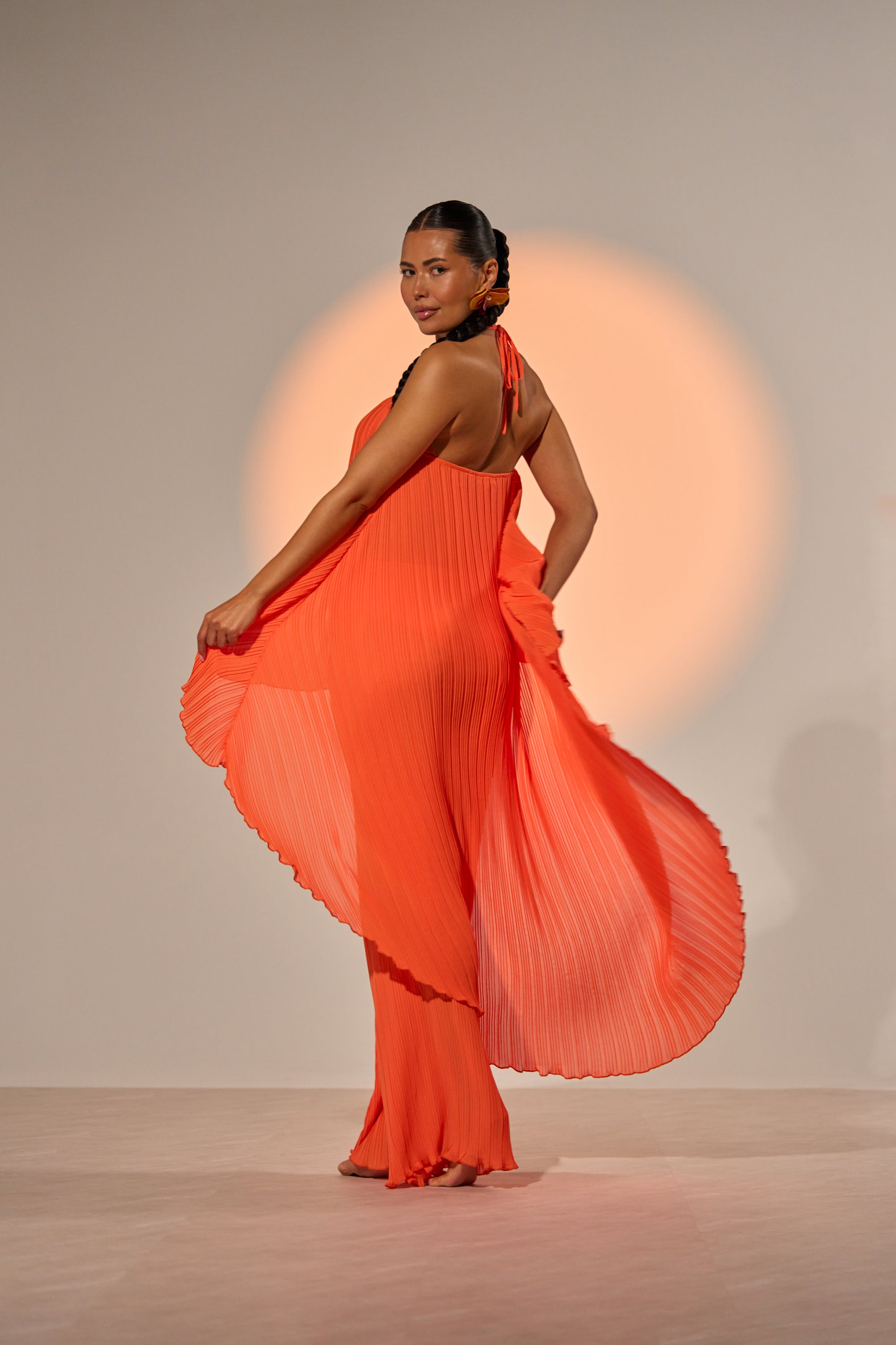 A woman poses gracefully on a light floor, wearing the CRUSHIN ON YOU BLOUSE—a bright orange halter styled as a pleated dress—looking over her shoulder with a gentle smile against a plain, warmly lit backdrop. 2