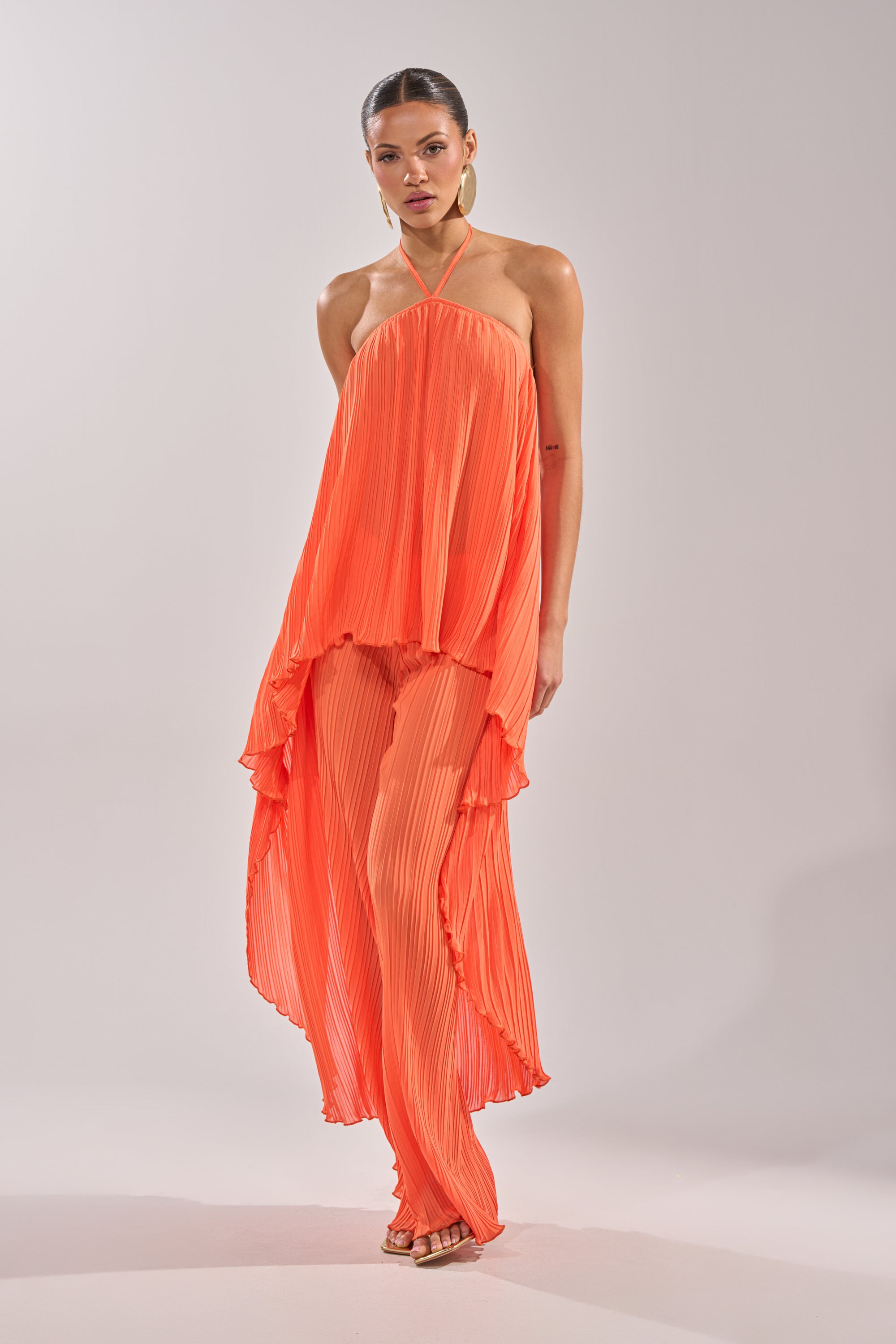 A woman wears the CRUSHIN ON YOU BLOUSE—bright orange, pleated, with a halter neckline and asymmetrical hem—paired with matching wide-leg pants and open-toe heels against a simple light background. 6