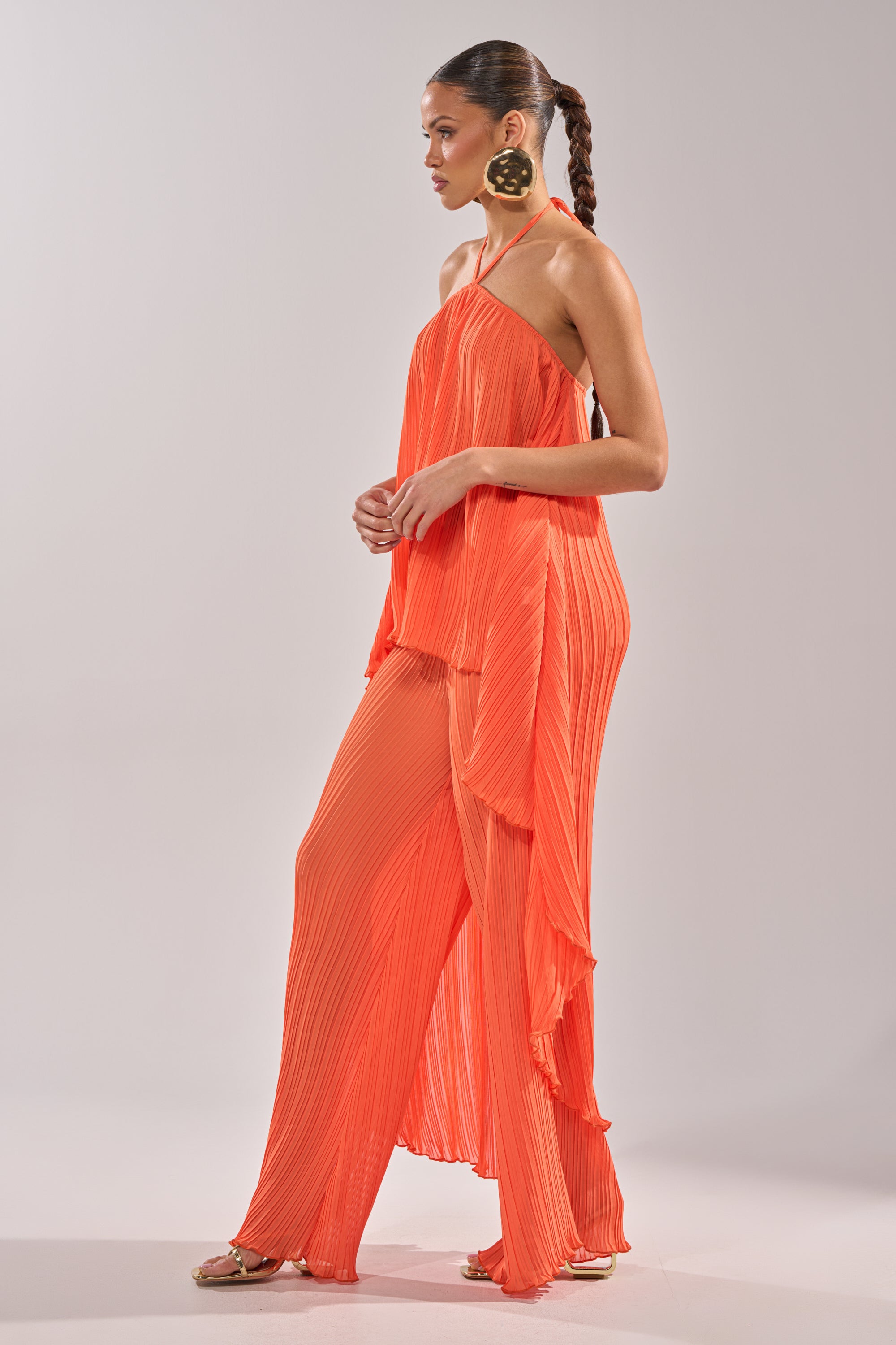 A woman stands in profile wearing the CRUSHIN ON YOU BLOUSE in sunset orange with matching wide-leg pants. She has a braided ponytail, statement earrings, and gold sandals, posed against a plain light background. 9