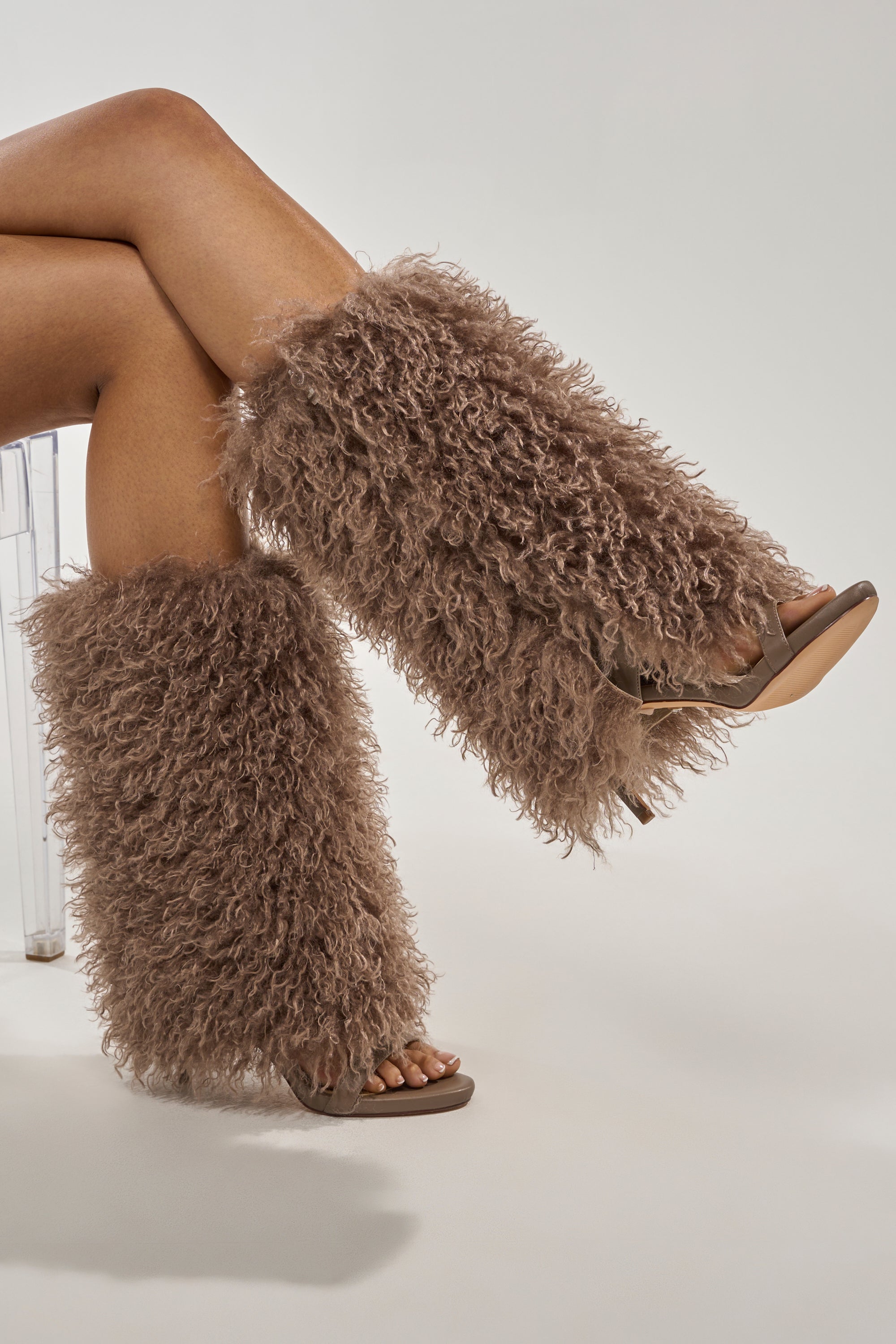 A person sits cross-legged against a plain light background, wearing the Azalea Wang CLOUD NINE Taupe Fur Open Toe Boot—knee-high sandal boots covered in thick, shaggy brown faux fur for a bold statement look. 1