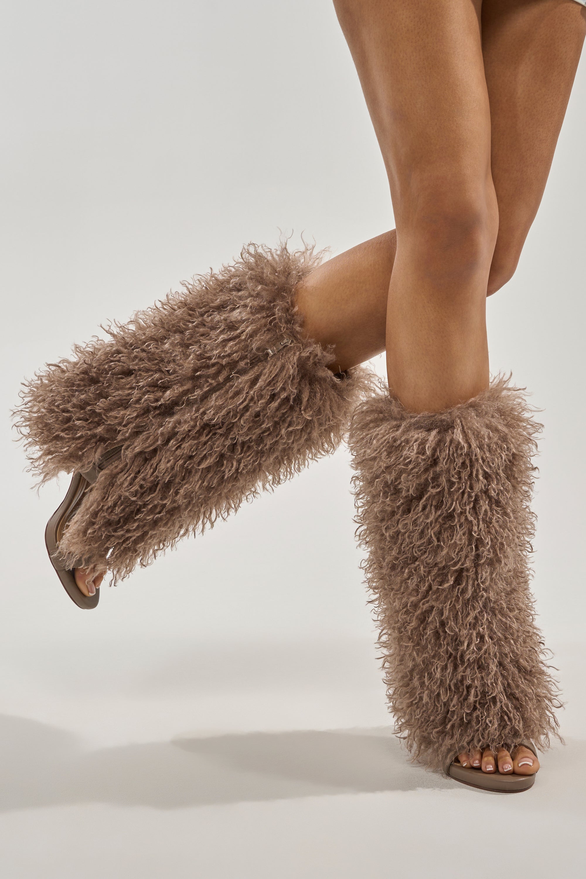 A person models the Azalea Wang CLOUD NINE TAUPE FUR OPEN TOE BOOT, posing with one foot lifted against a plain light background. 7