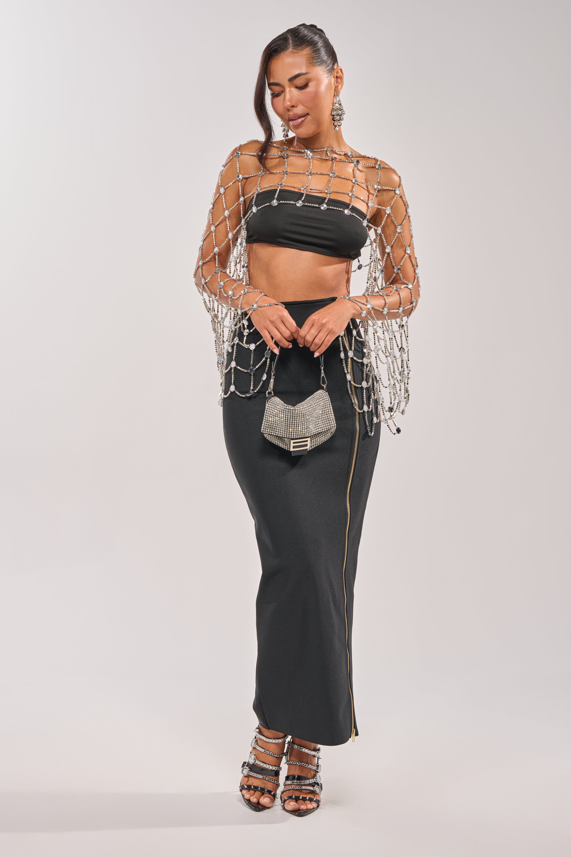 A woman wears the CHAINED TO YOU TOP with a long black skirt, a silver netted shawl with metallic accents, strappy heels, and holds a small silver handbag against a plain background. 10