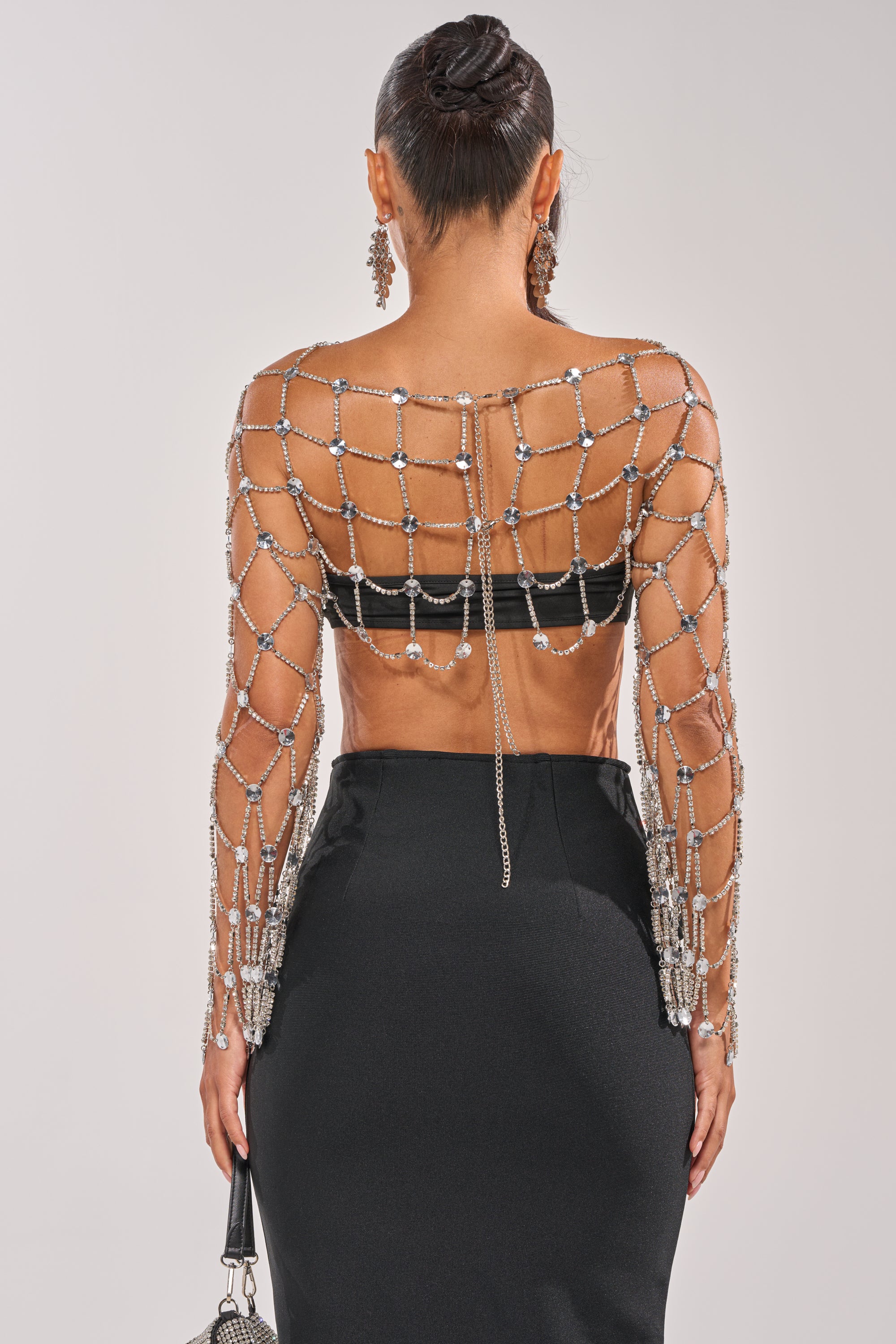 A woman with her back to the camera wears the CHAINED TO YOU TOP—a rhinestone lattice design with sheer long sleeves—paired with a black skirt and neat bun, carrying a small handbag for the perfect festival look. 6