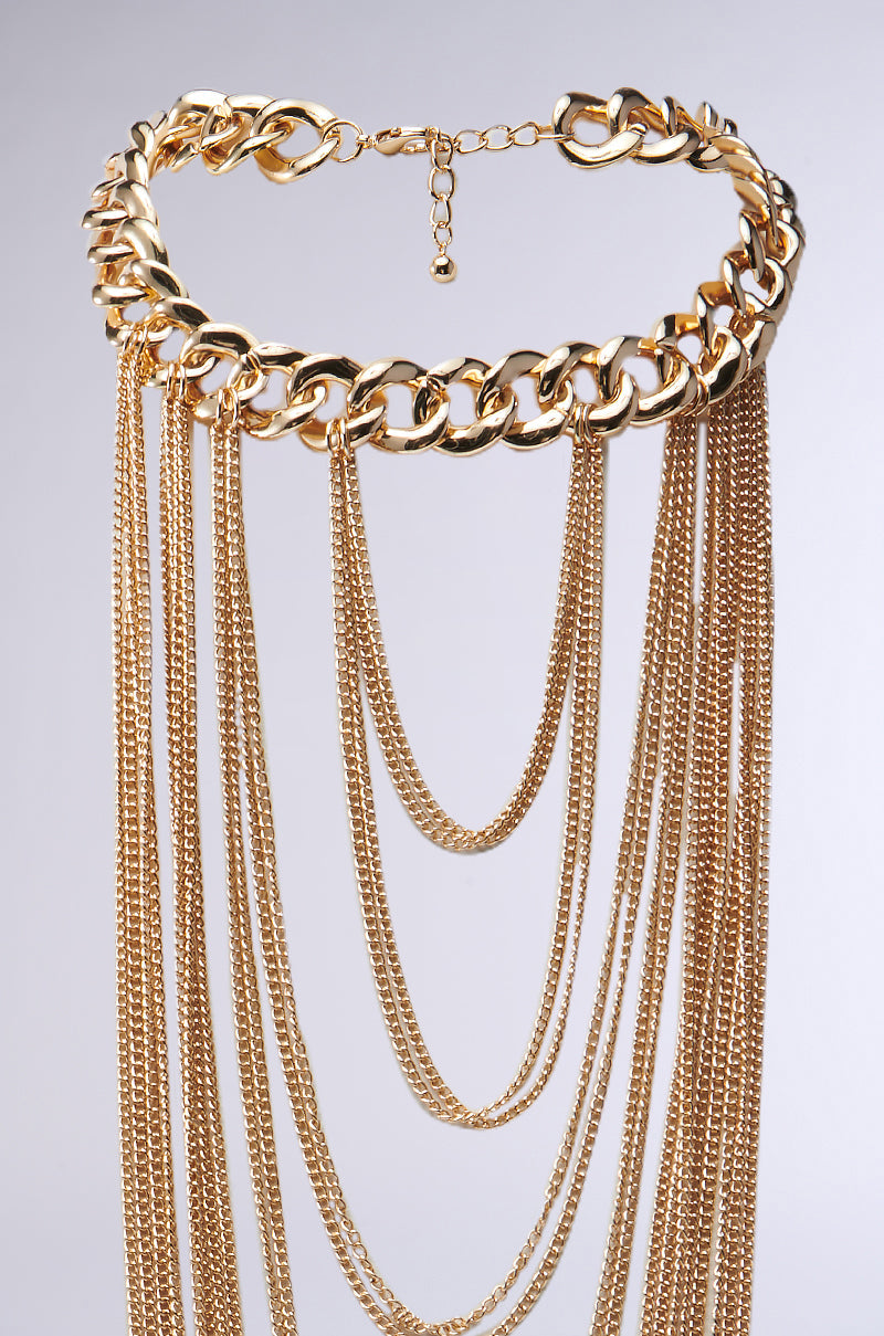 Introducing the SO MANY LAYERS NECKLACE, a statement piece with a multilayered look. It features thick gold chains alongside cascading layers of thinner ones, creating a tiered effect that adds depth and texture against a plain gray background. 3