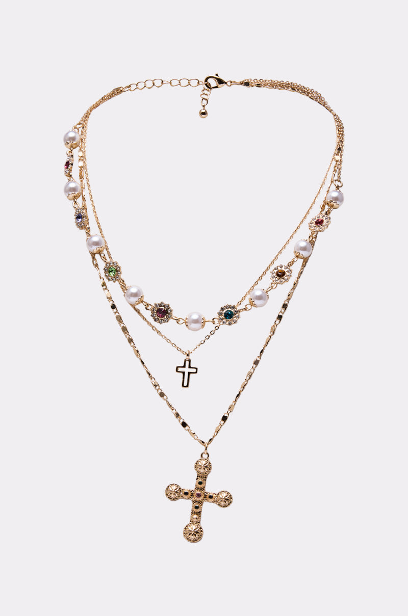 The LIKE A PRAYER GOLD CROSS NECKLACE features layered gold-tone chains with faux pearls, colorful gemstones, a black cross charm, and an ornate gold cross pendant—creating a bold statement jewelry piece.
