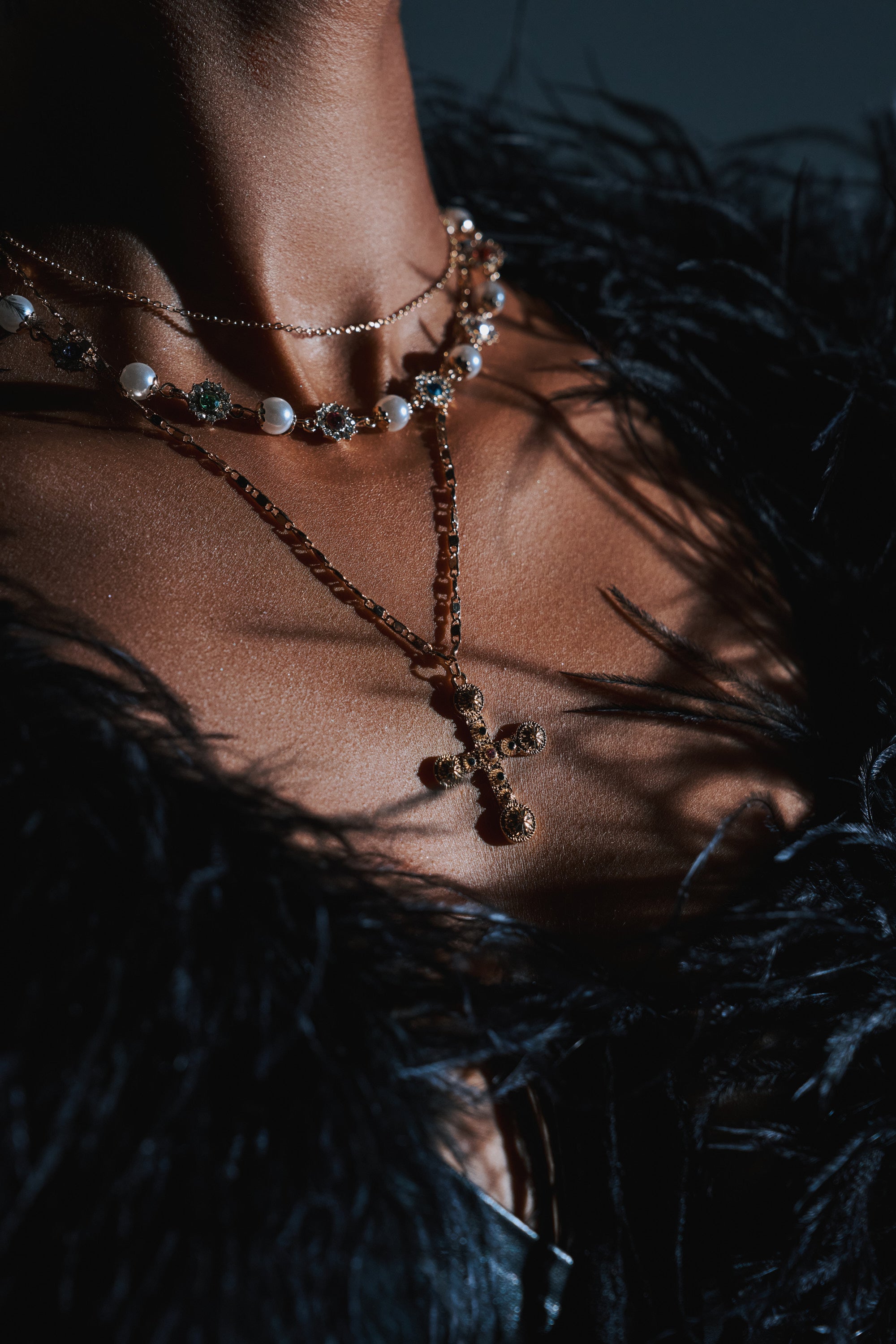 A close-up of a neck adorned with the LIKE A PRAYER GOLD CROSS NECKLACE, layered with pearls above a black feathery top. Dramatic lighting accentuates the statement jewelry, casting striking shadows on the skin.