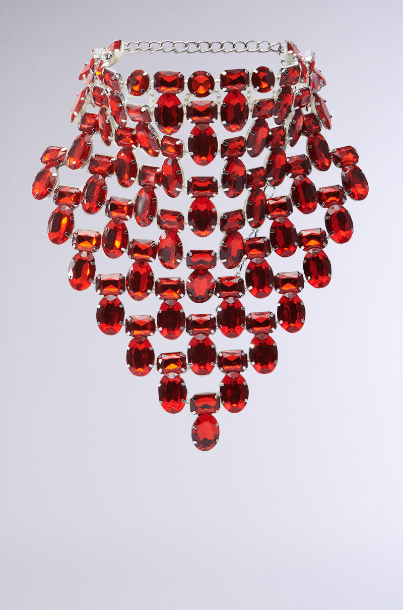 The ATTRACTING WEALTH STATEMENT NECKLACE features cascading rows of ruby-inspired jewels set in faux silver, with large faceted oval gemstones creating a bold triangular silhouette.