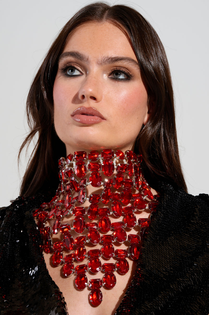 A woman with long brown hair and dramatic makeup wears the ATTRACTING WEALTH STATEMENT NECKLACE, adorned with ruby-inspired jewels covering her neck and upper chest. She’s dressed in a black sequined outfit, gazing slightly upward.