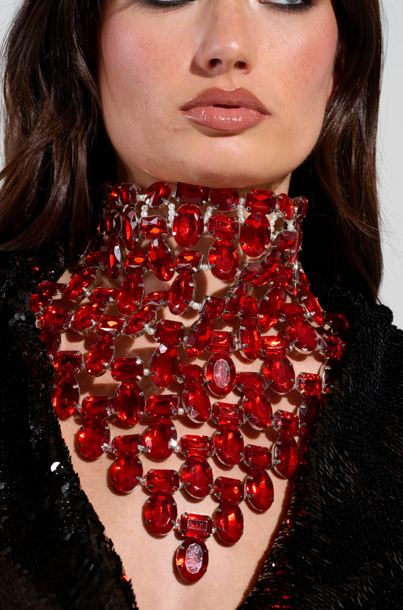 A woman wears the ATTRACTING WEALTH STATEMENT NECKLACE, featuring large, oval ruby-inspired jewels in faux silver metallic, paired with a black sequined outfit. She has glossy lips and subtle makeup.