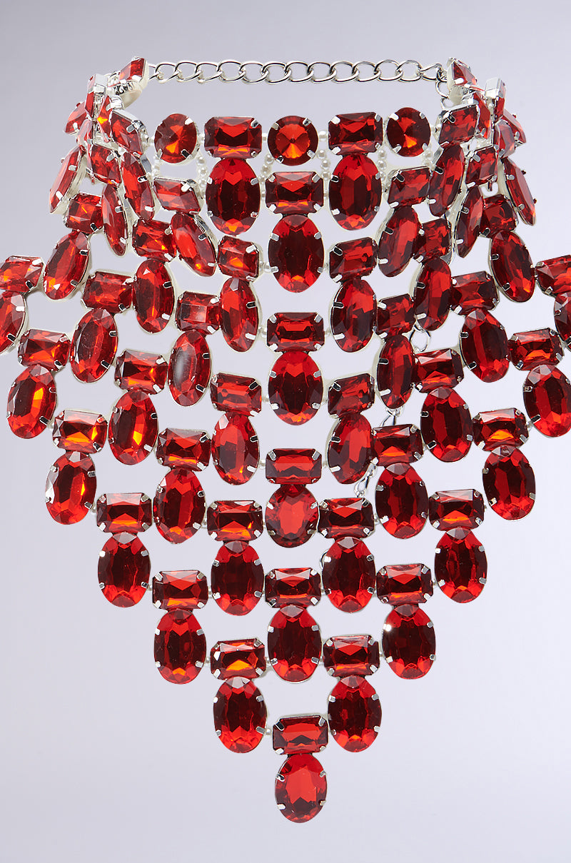 The ATTRACTING WEALTH STATEMENT NECKLACE showcases cascading rows of large, ruby-inspired jewels set in faux silver metallic, arranged in a striking triangular pattern.