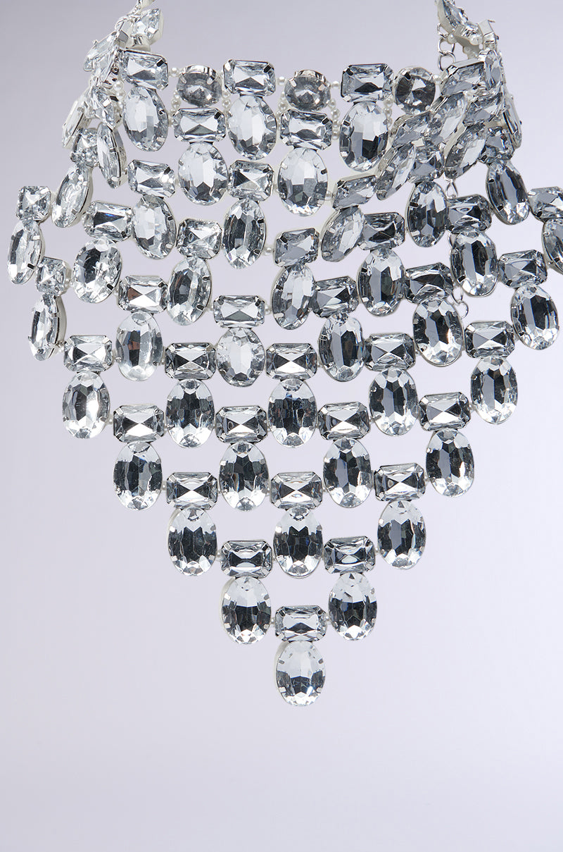 A luxurious chandelier with multiple rows of teardrop-shaped glass crystals, arranged in a cascading pattern, reflects light to create a sparkling effect against a soft gray background, capturing the elegance reminiscent of the PAY MY BILLS NECKLACE IN SILVER.