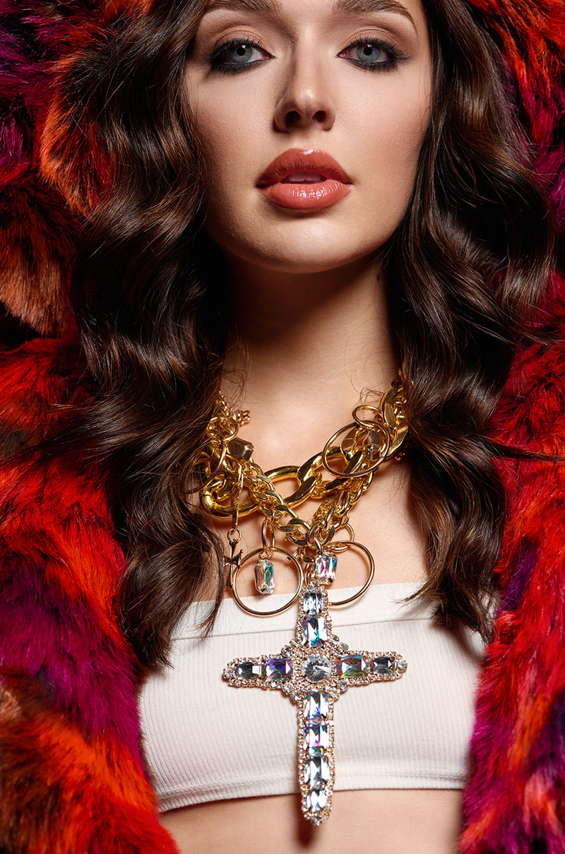 A woman with wavy hair wears a striking furry red and orange coat over a white top. Against the dark backdrop, her LIKE A VIRGIN STATEMENT NECKLACE gleams—a stack of layered gold chains featuring a large jeweled cross pendant that captures the eye.