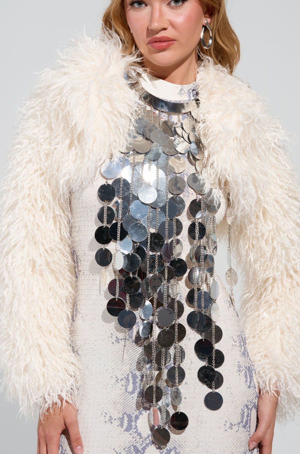A person is dressed in a white, furry jacket over a white dress decorated with cascading silver sequin discs, complemented by the INVITE ME TO THE PARTY STATEMENT NECKLACE. With light brown hair and posed against a neutral background, they exhibit a chic and contemporary fashion ensemble.