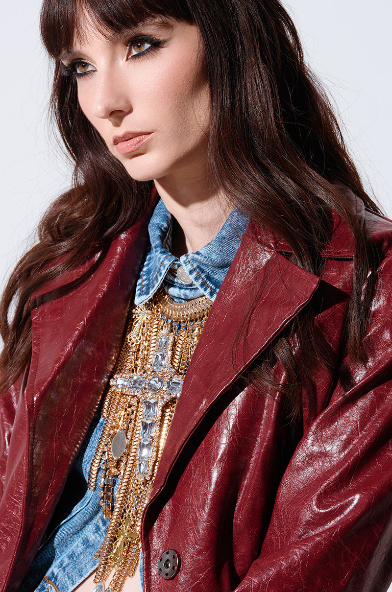 A woman with long, wavy brown hair is wearing a blue denim shirt beneath a shiny red leather jacket. She sports the CROSS MY HEART CHAIN NECKLACE, which features metallic gold links and an adorned cross pendant. The background is plain and light-colored.