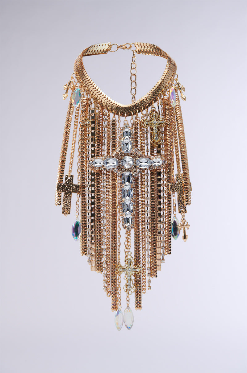 The CROSS MY HEART CHAIN NECKLACE is a striking jewelry piece featuring multiple cascading gold chains adorned with various crystals and cross pendants. The centerpiece boasts a large crystal cross, while the surrounding gold chains are decorated with smaller crystals and cross charms, making this necklace an exquisite statement accessory.