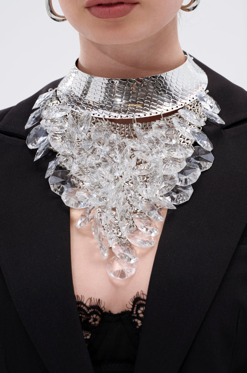 A person wearing the PARTY STARTER LUXE NECKLACE featuring crystal-clear gems and a wide, textured metallic choker, styled with a black blazer and lace-trimmed top. 1