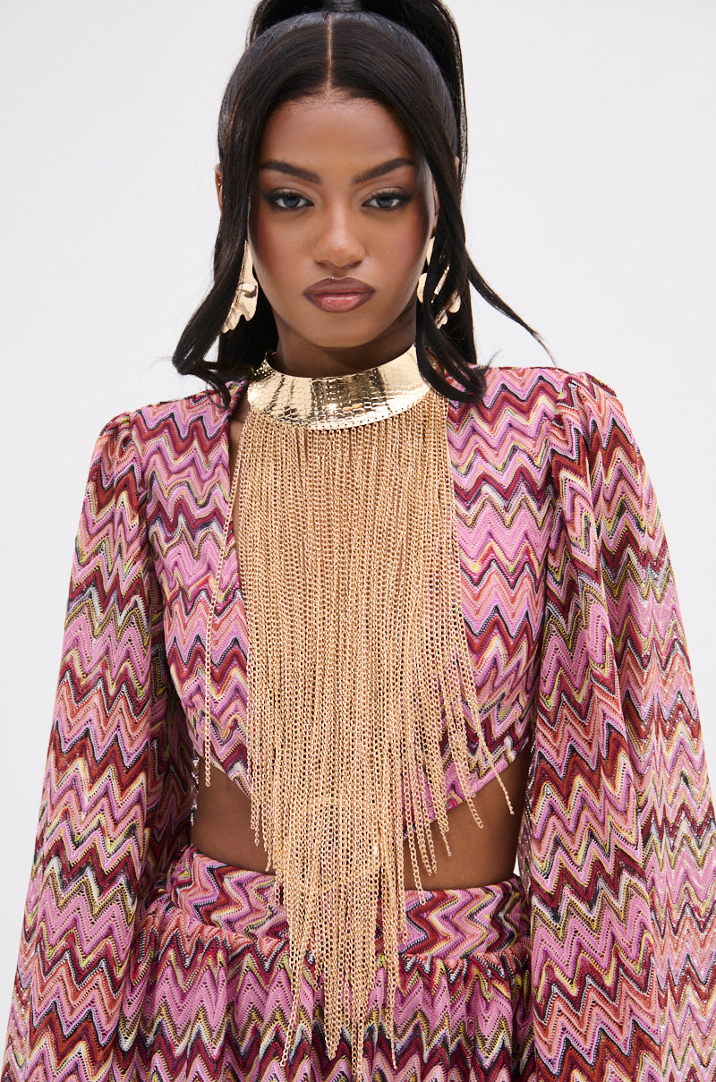 A person wearing a vibrant zigzag-patterned outfit with bell sleeves, a matching headpiece, and a STAY DRIPPIN CHAIN NECKLACE has long dark hair in a partial updo against a plain white background.
