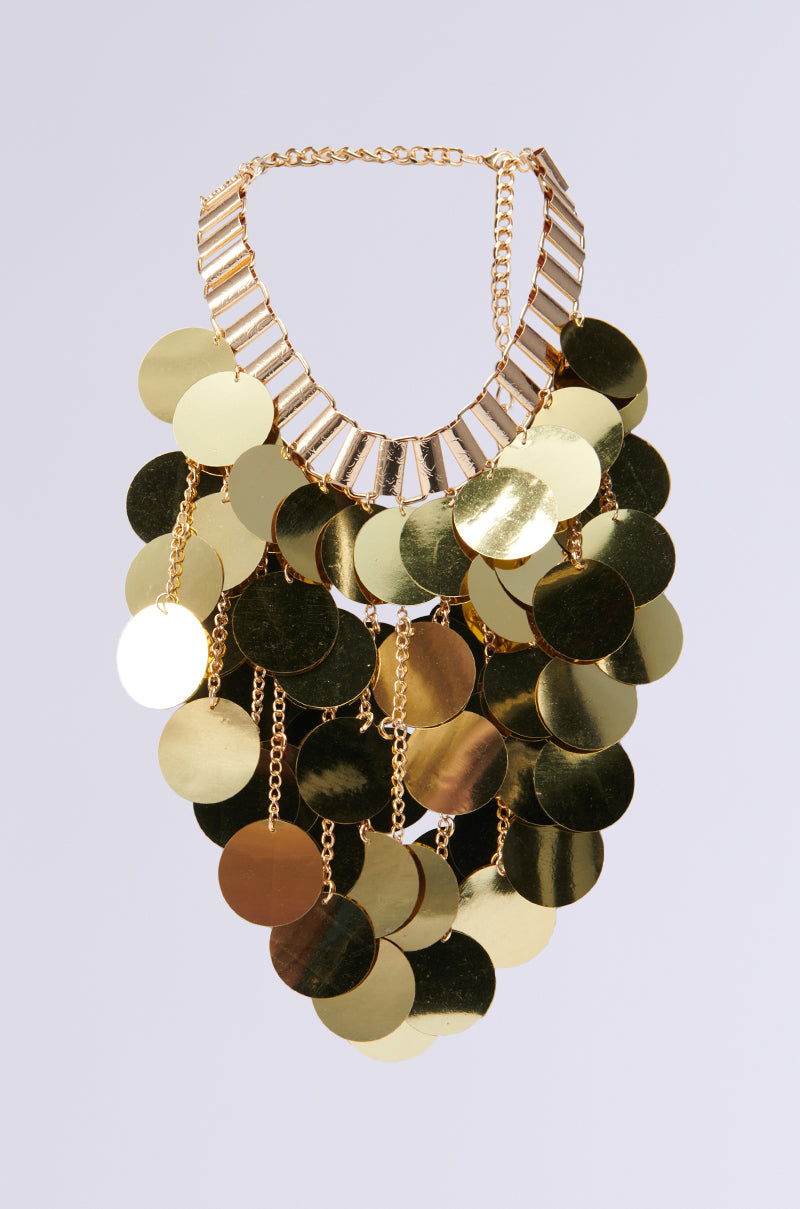 The ALL NIGHT LONG SEQUIN NECKLACE is a captivating party accessory, showcasing large, round metallic gold discs arranged in tiers. These discs are elegantly affixed to a gold chain with rectangular links, creating a dazzling, cascading appearance against the simple gray backdrop.