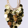 The ALL NIGHT LONG SEQUIN NECKLACE is a captivating party accessory, showcasing large, round metallic gold discs arranged in tiers. These discs are elegantly affixed to a gold chain with rectangular links, creating a dazzling, cascading appearance against the simple gray backdrop.