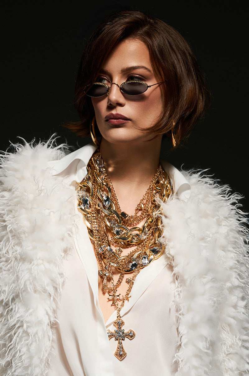 A woman with short hair wears small round sunglasses and the CROSS ME NECKLACE IN GOLD, a chunky multi-layered accessory featuring a cross charm, complemented by large hoop earrings. She is dressed in a white feathered coat over a white shirt, set against a dark background.