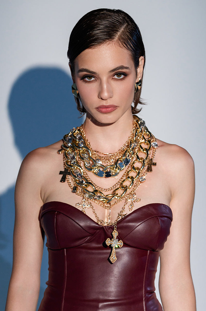 A woman with short, slicked-back hair wears a strapless burgundy leather dress and the CROSS ME NECKLACE IN GOLD—layered chunky gold chains with cross pendants—posing against a plain background with a soft shadow.