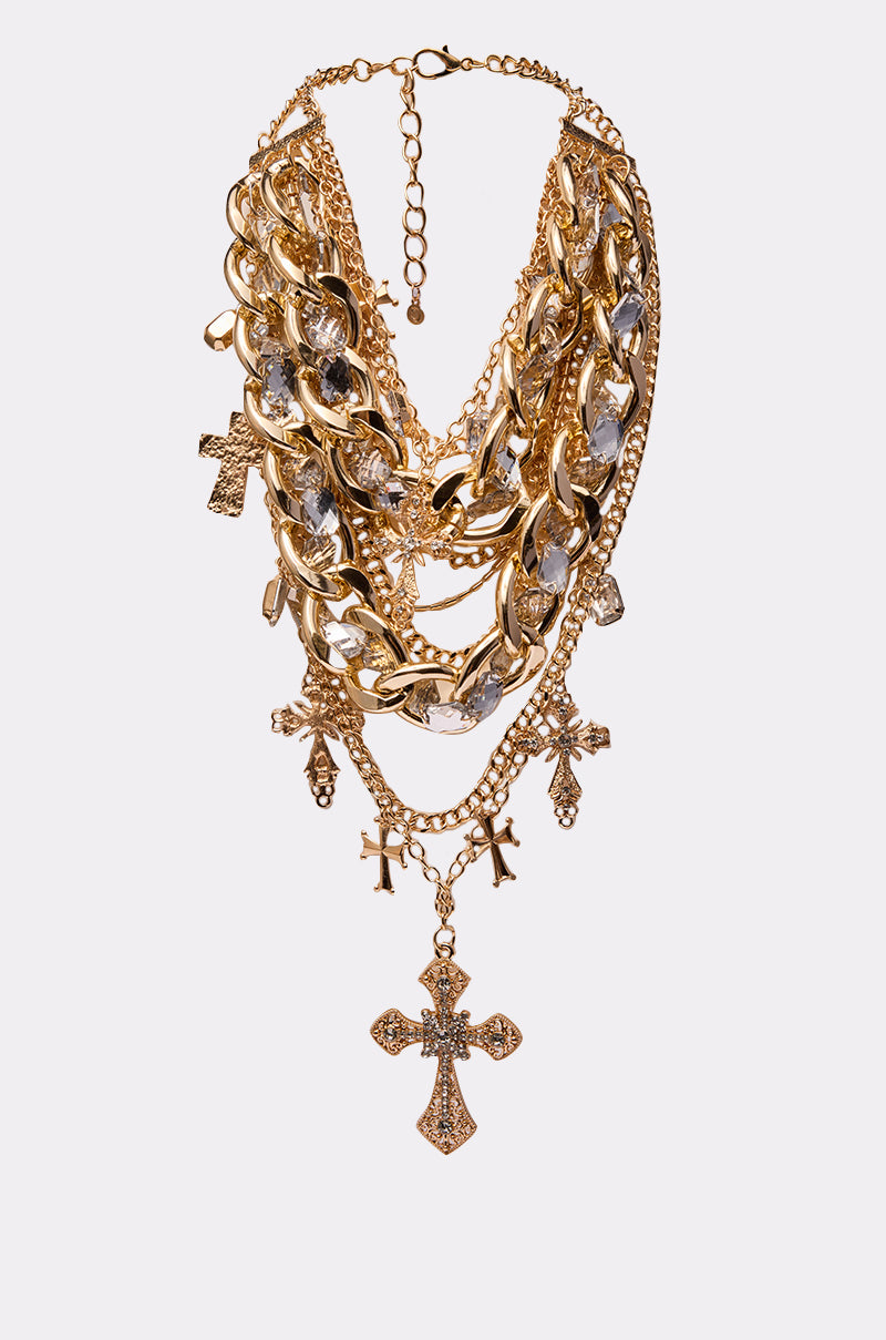 The CROSS ME NECKLACE IN GOLD features layered thick gold chains with ornate cross pendants adorned with clear gemstones, shown on a white background.