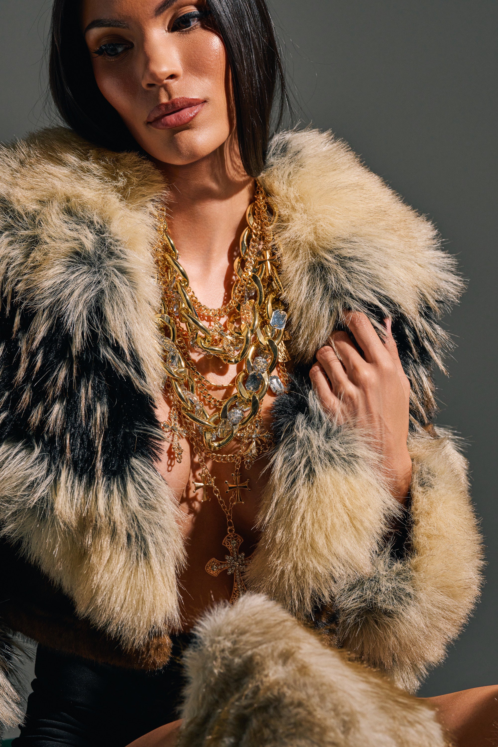 A woman with straight dark hair and a neutral expression stands against a plain gray background, wearing the CROSS ME NECKLACE IN GOLD—a chunky gold necklace layered with a cross pendant—and a luxurious fur coat.