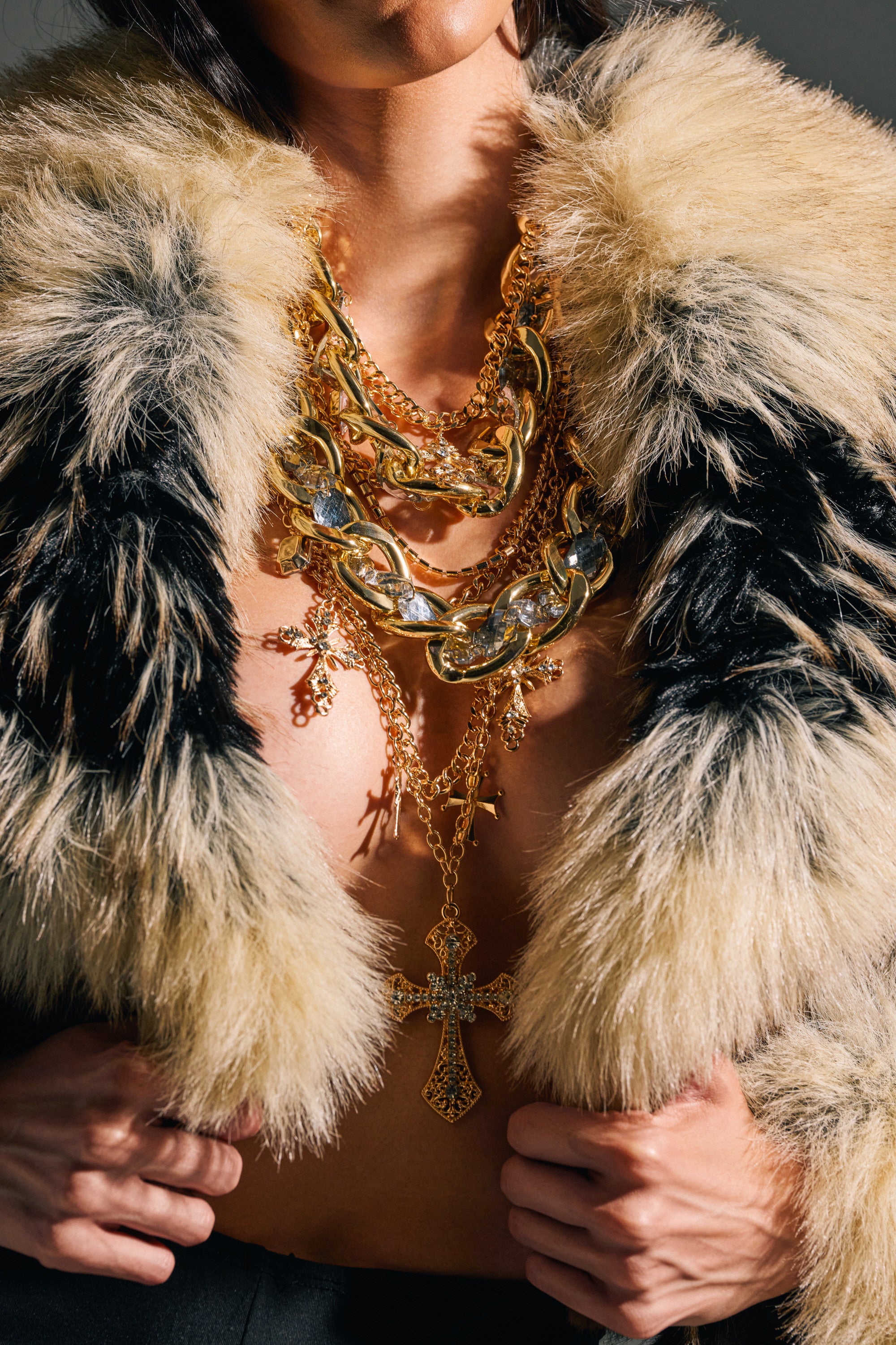 A person wears a fluffy black and beige fur coat, styled open at the chest to showcase the CROSS ME NECKLACE IN GOLD—layered with chunky chains and a bold jeweled pendant for a striking statement look.