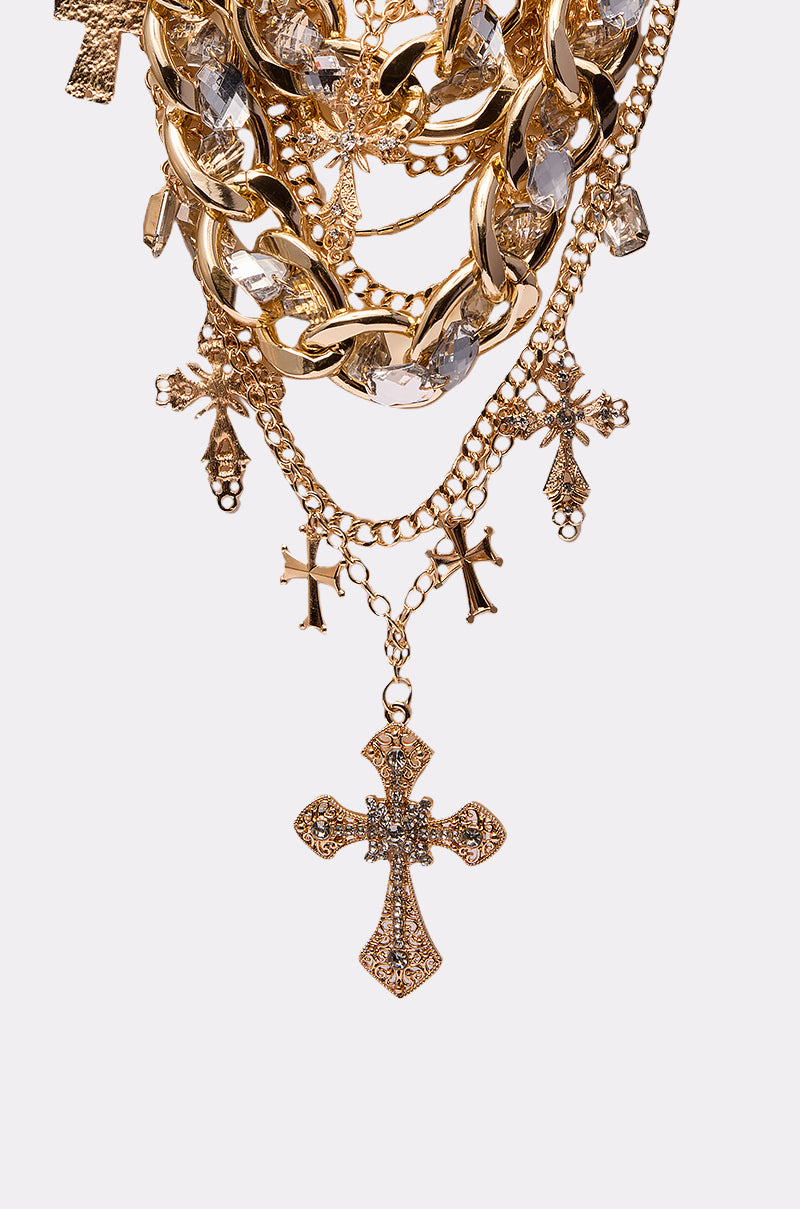 The CROSS ME NECKLACE IN GOLD features ornate cross pendants with clear gemstones on both small and chunky gold chains, creating a stunning layered look against a plain white background.