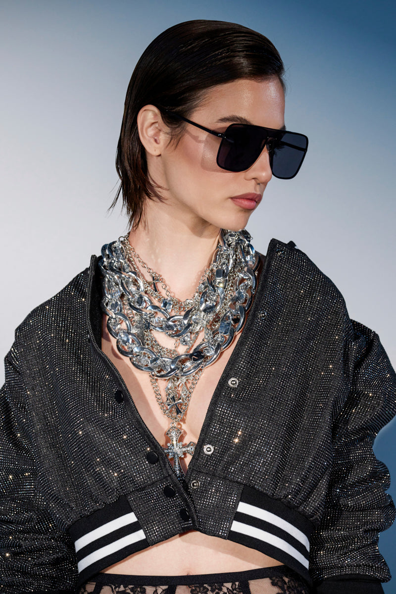 Wearing oversized black sunglasses and a sparkly black jacket with black lace underneath, a person with slicked-back dark hair shows off the CROSS ME METAL NECKLACE featuring a rhinestone cross pendant against a gradient blue background.