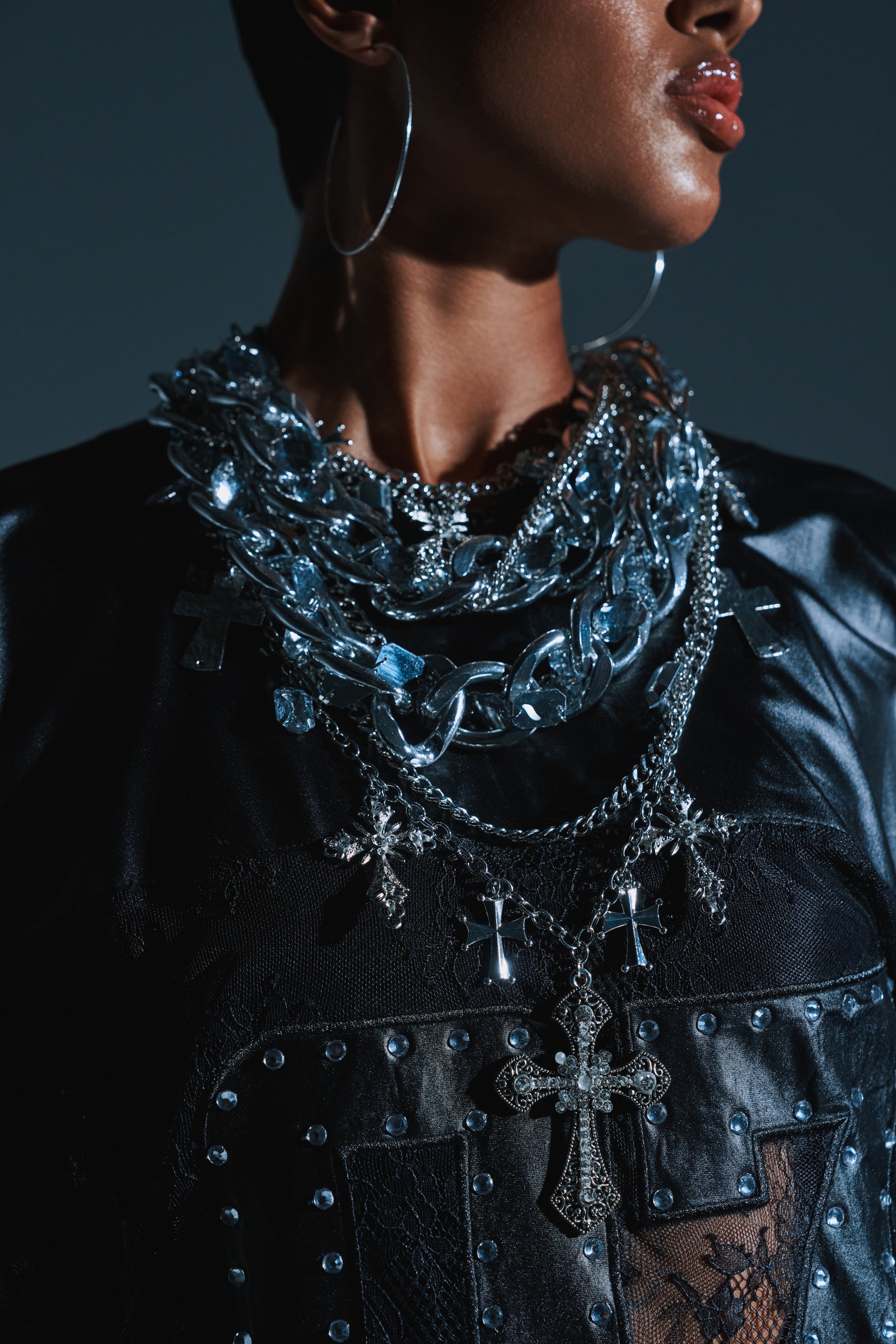 A person wearing large silver hoop earrings, the CROSS ME METAL NECKLACE layered with silver chains, and a black leather top with metal studs and lace details.