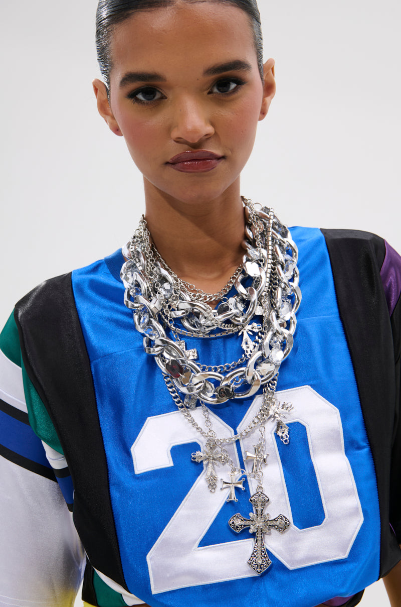 A woman with sleek, dark hair wears a blue jersey numbered 20 and the CROSS ME METAL NECKLACE—layered chunky chains with a cross pendant—standing against a plain white background.