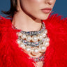A woman in a vibrant red feathered outfit wears the PRETTY IN PEARLS NECKLACE—a bold, layered pearl and crystal piece—as she gazes to the side against a blue background.
