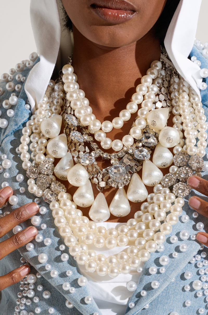 A close-up of a person wearing a PEARL PLEASE STATEMENT NECKLACE, flaunting numerous pearl and rhinestone necklaces of various sizes alongside large jeweled embellishments. The jacket, adorned with pearls, complements the overall lavish pearl-encrusted theme. 1