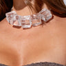 A woman wearing the ICED OUT CHOKER NECKLACE, which features large, clear, cube-shaped beads. She is dressed in a textured, dark-colored strapless top, and her long brown hair cascades effortlessly down her shoulders.