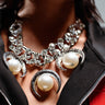 Close-up of a person's neck adorned with the WESTERN VIBEZ NECKLACE, showcasing large pearl accents encased in silver claw-like settings. The necklace is composed of interlocking silver rings. The person wears a black jacket with a red detail.