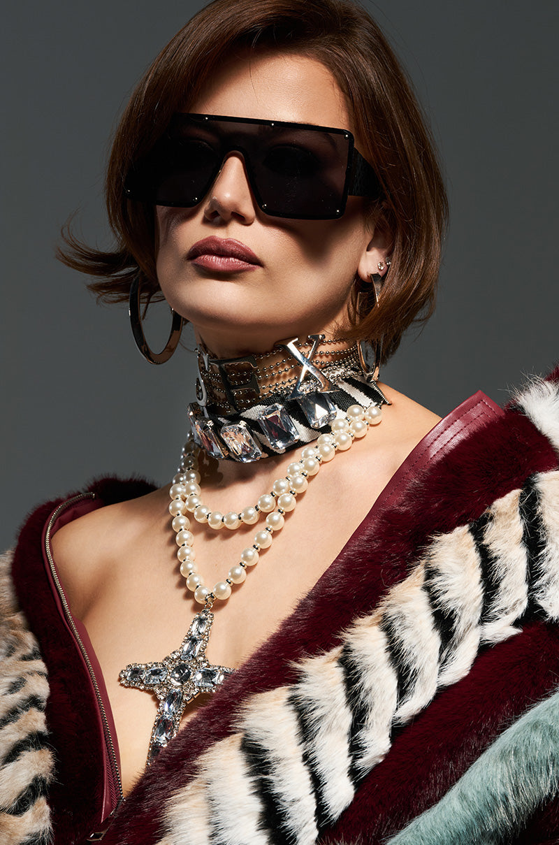 The individual is adorned with large dark sunglasses, hoop earrings, and layered necklaces featuring the WALK ON THE WILD SIDE NECKLACE among them. They sport a short hairstyle and are elegantly dressed in a luxurious fur coat showcasing bold zebra patterns and subtle gemstone embellishments.