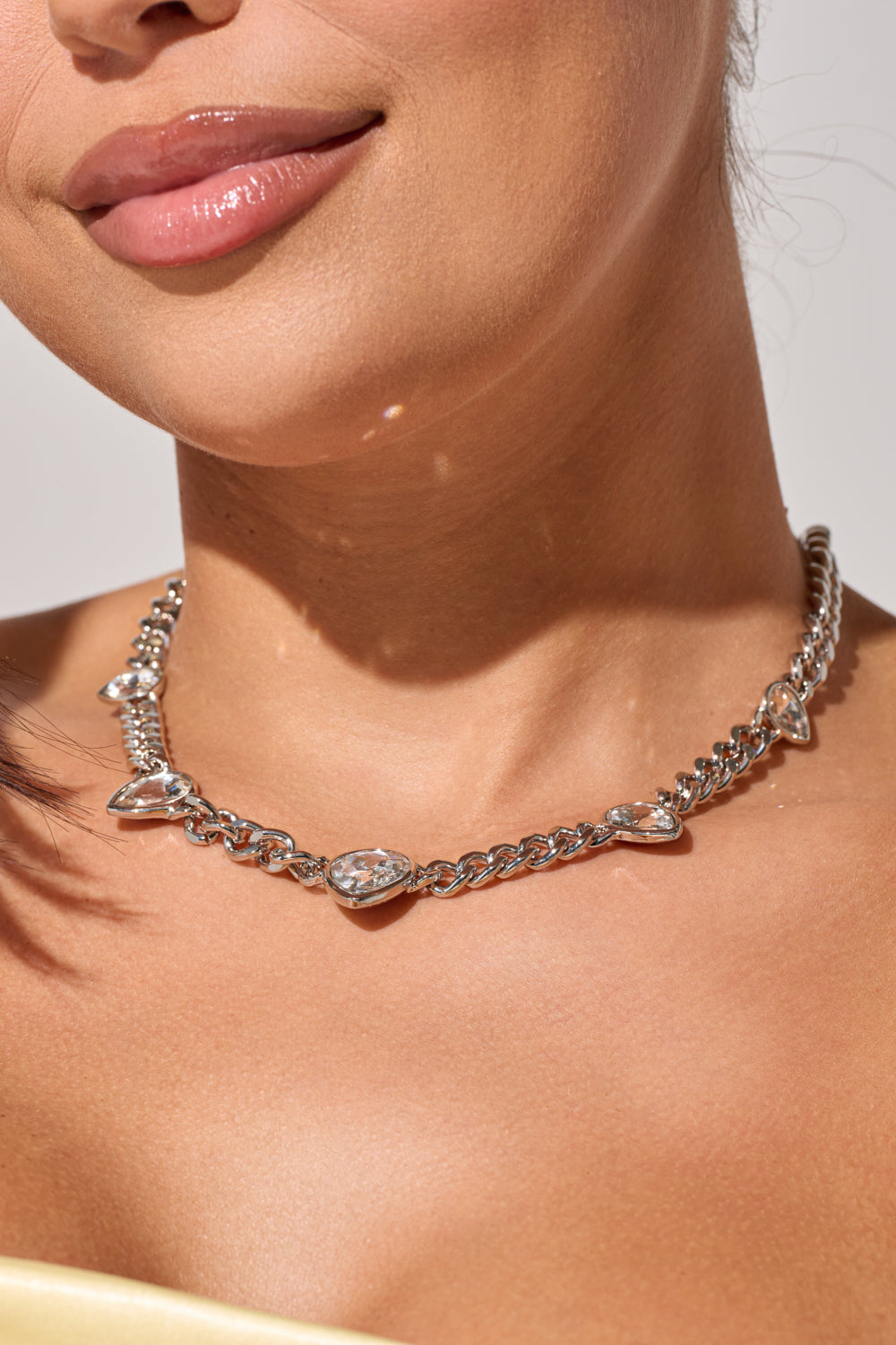A close-up of a woman wearing the BESS NECKLACE, featuring silver heart-shaped pendants and subtle rhinestone accents. She has smooth skin, glossy lips, and is dressed in a pale yellow top. 0