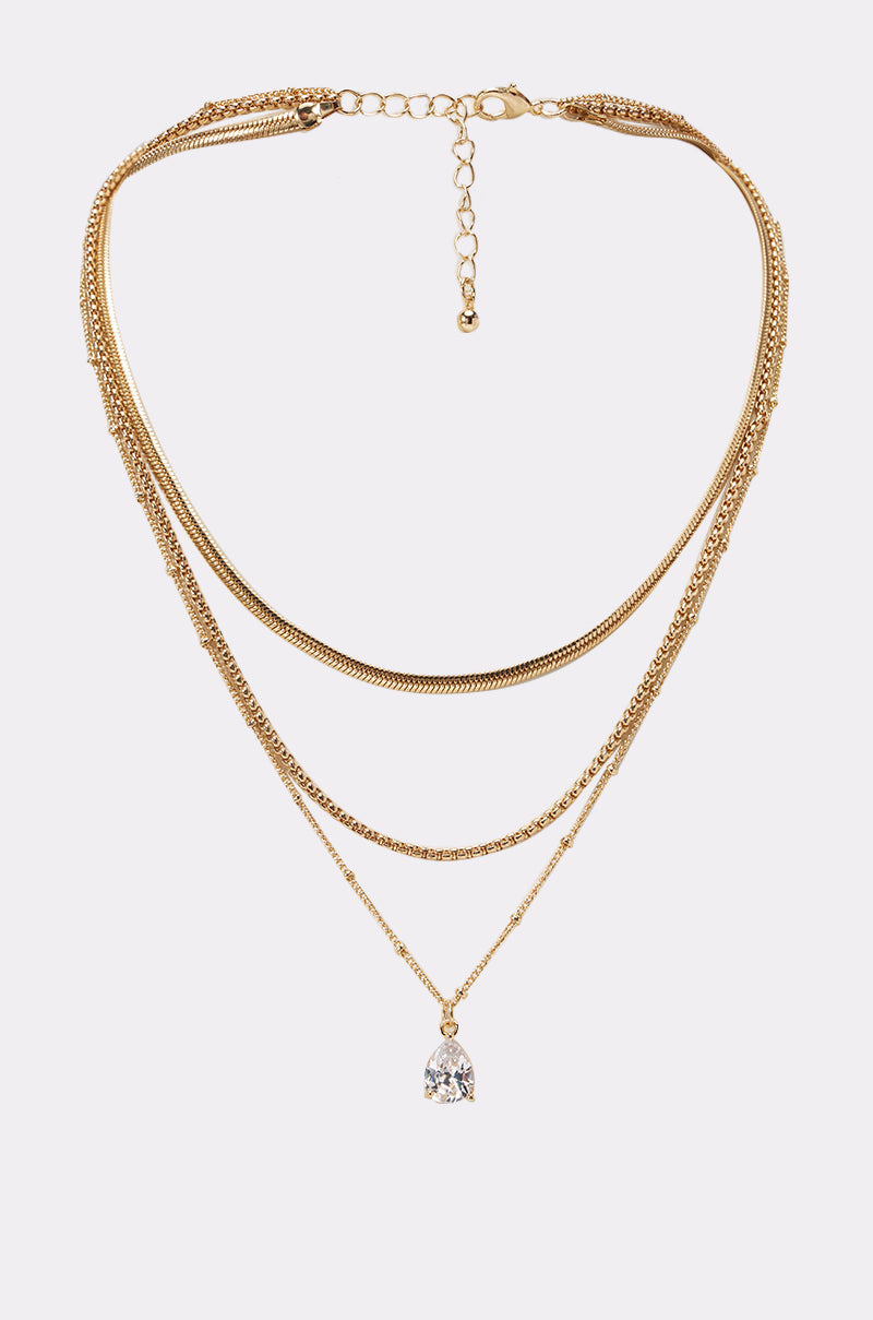 The COMPLETE THE LOOK NECKLACE features four layered gold chains of varying thickness, with an adjustable fit; the shortest is a flat herringbone, and the longest displays a teardrop crystal pendant.